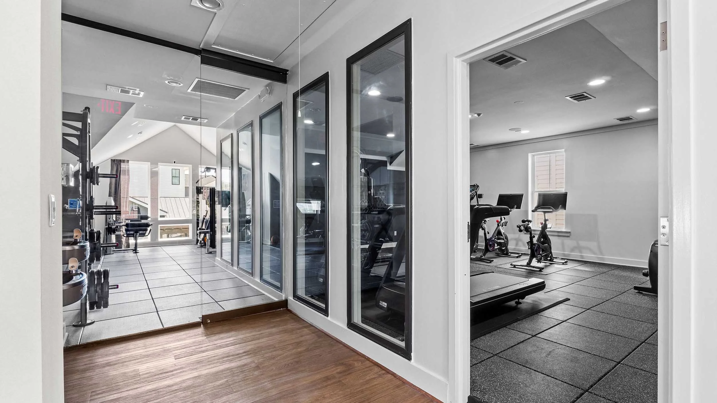 Fitness room with exercise equipment, including bikes, a treadmill, and weight machines, viewed through glass doors in a modern gym.