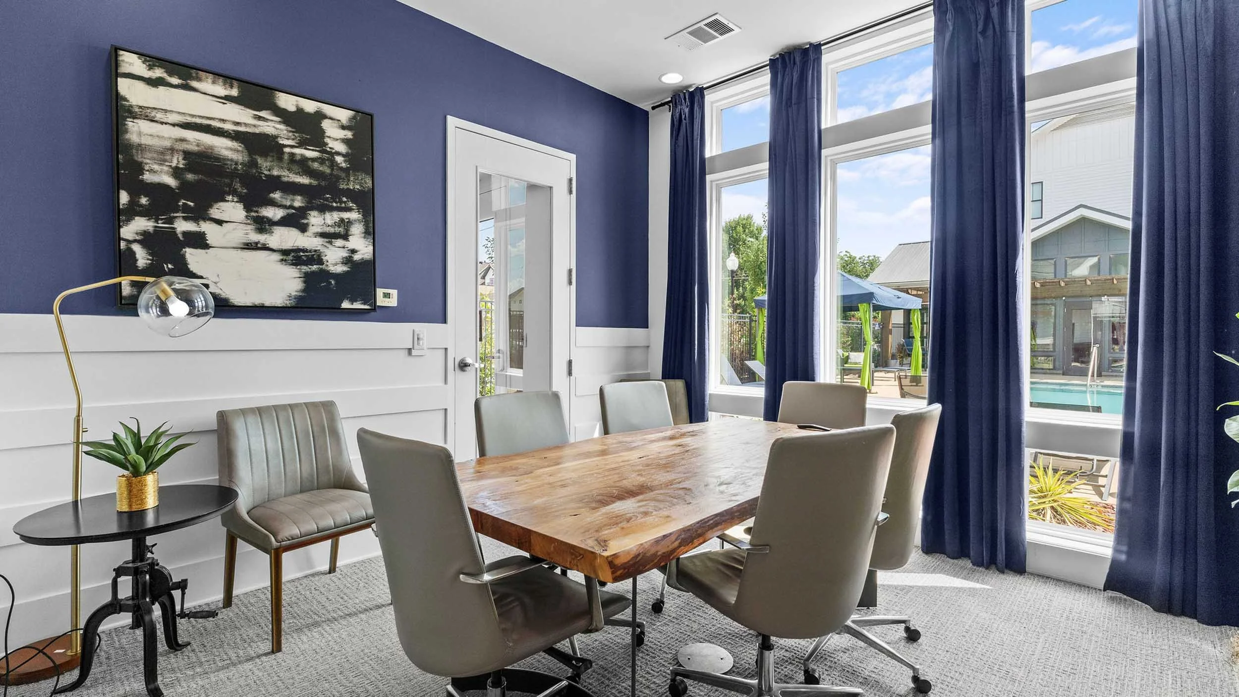 A study room with a wooden table and six gray chairs. Blue curtains frame large windows that overlook a backyard with patio furniture, a blue umbrella, and a pool. A black and white abstract painting hangs on a dark blue wall.