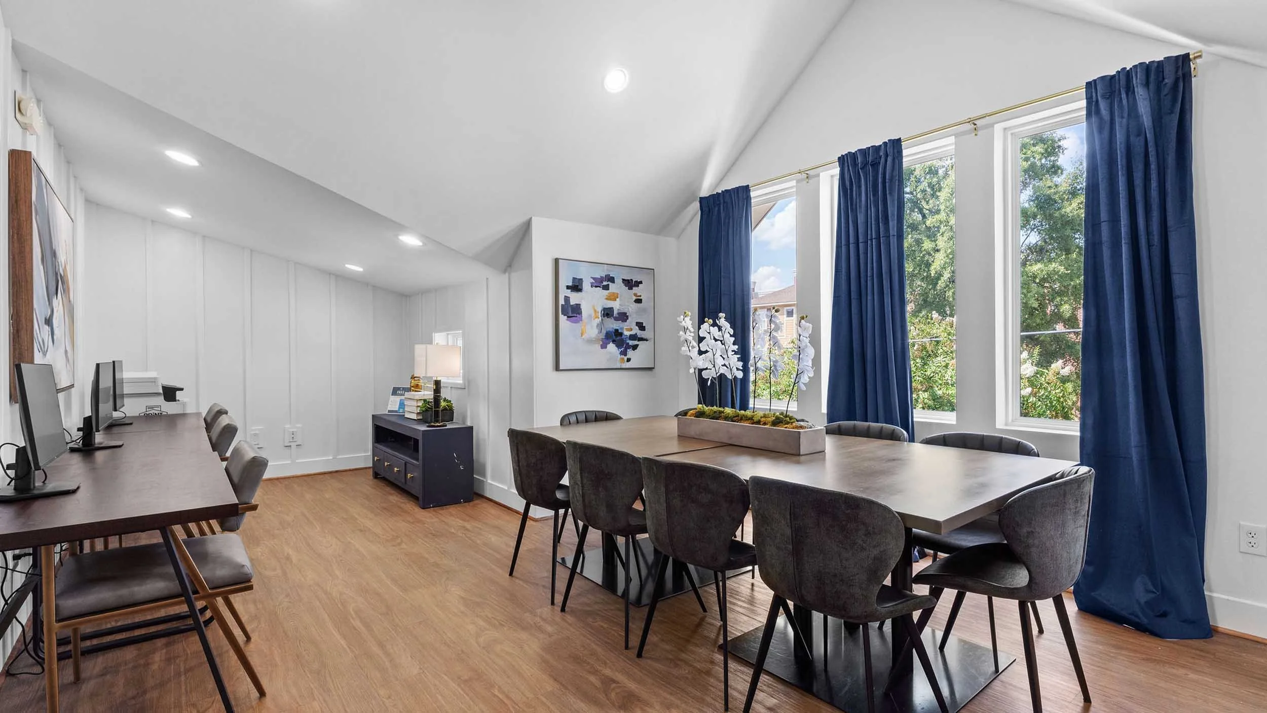 Office conference room with a long table, black chairs, large windows with blue curtains, and abstract artwork on white walls.