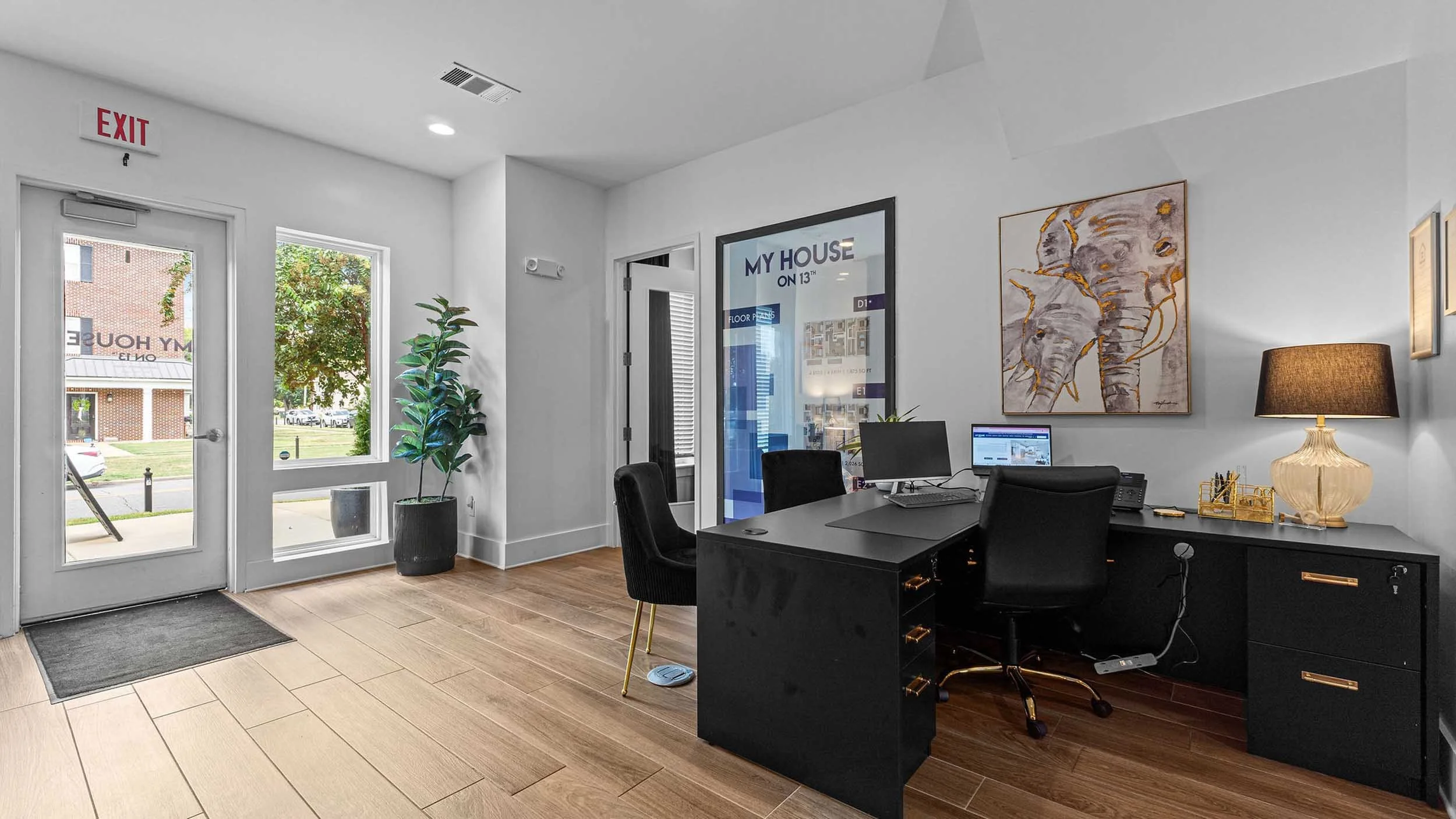 Office reception area with a black desk, computer, black chairs, a lamp, artwork, a potted plant, and windows and door leading outside.