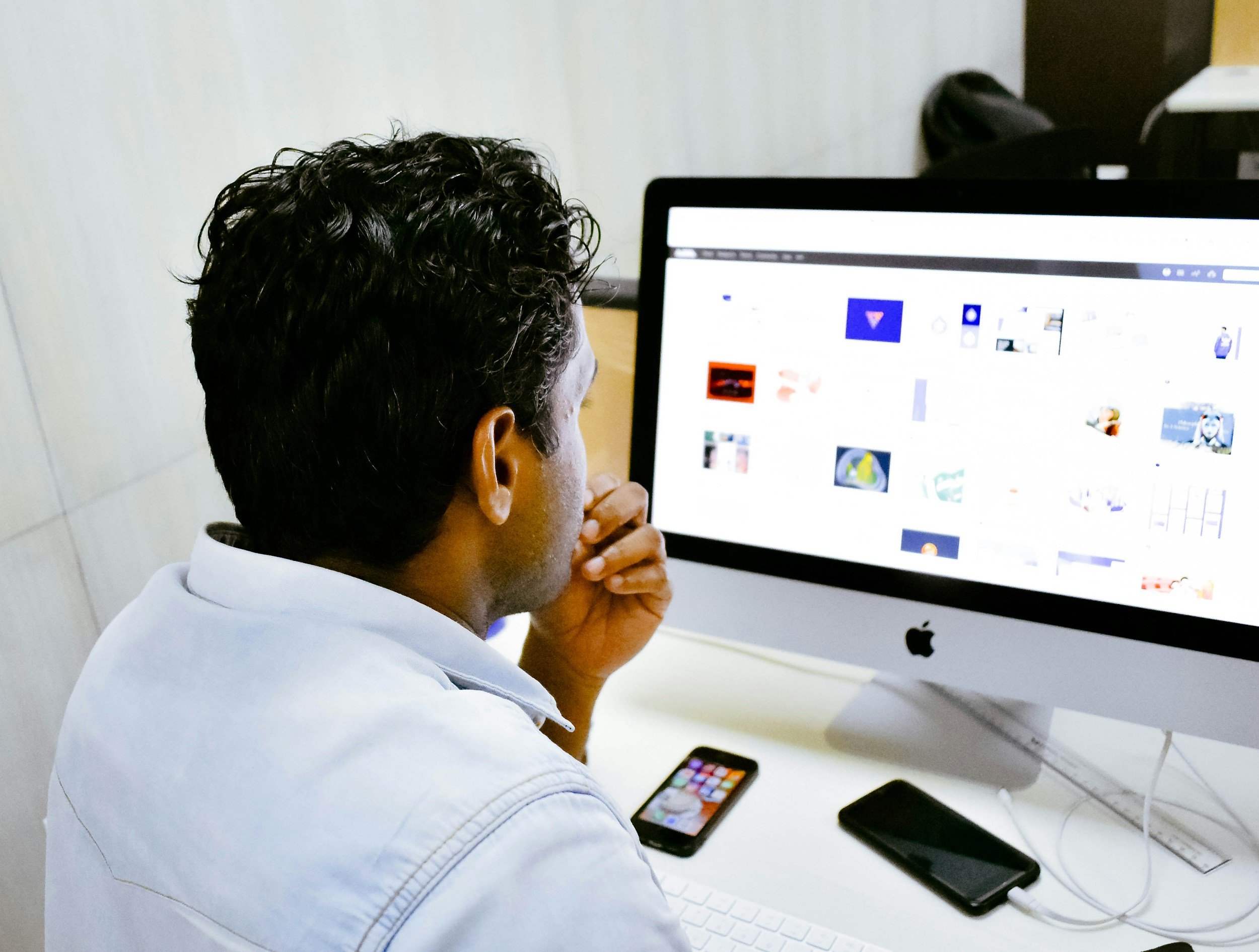 A man with curly black hair working on a computer with a large screen, an iPhone, and another smartphone nearby.