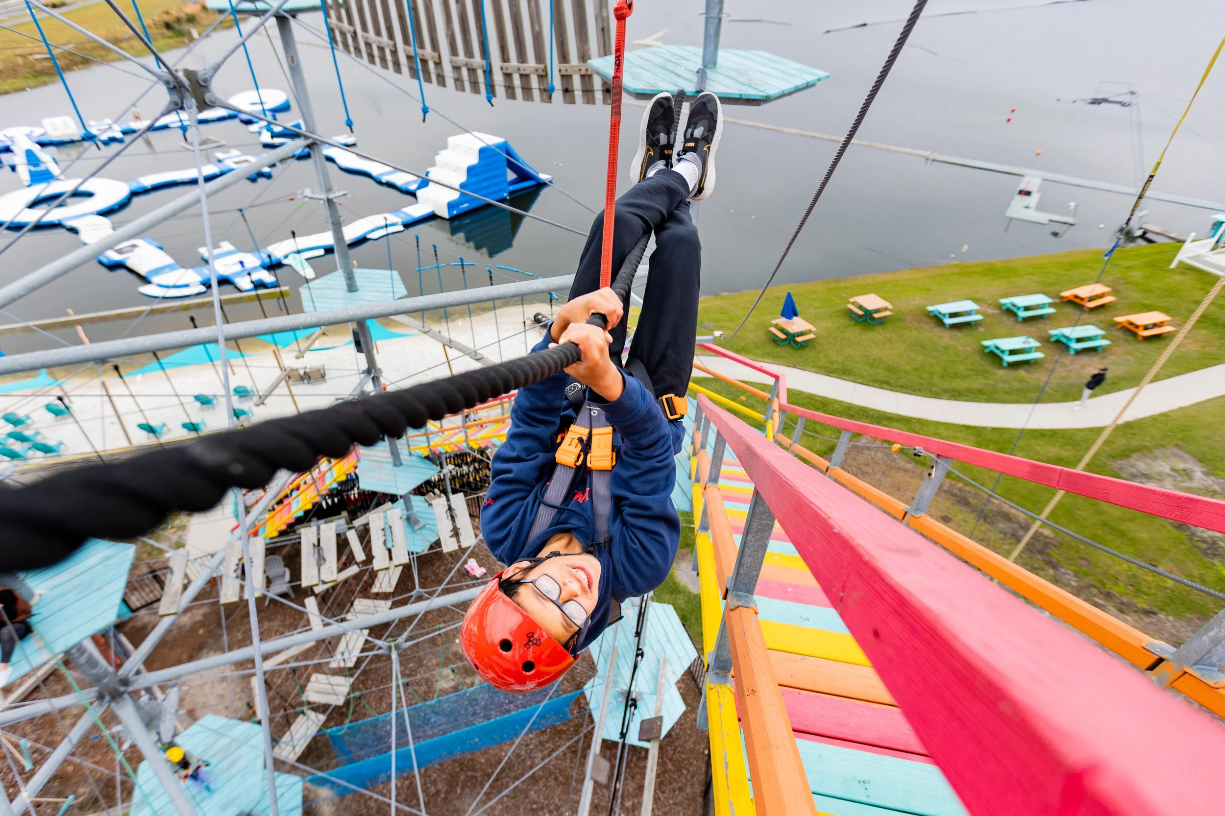 A teenager hanging upside down from a rope obstacle on an aerial ropes course with a beach and aqua park and picnic benches in the background.