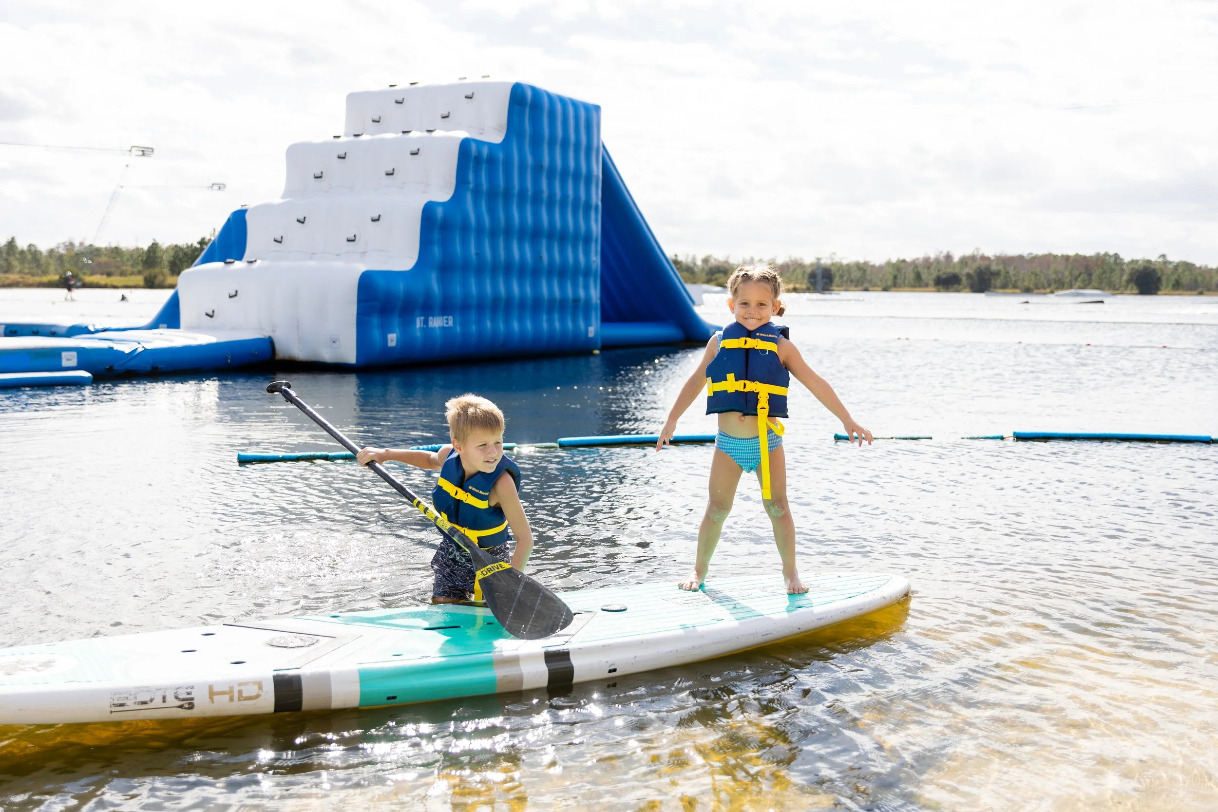 Two children wearing life jackets paddleboarding near a large inflatable water slide on a lake.