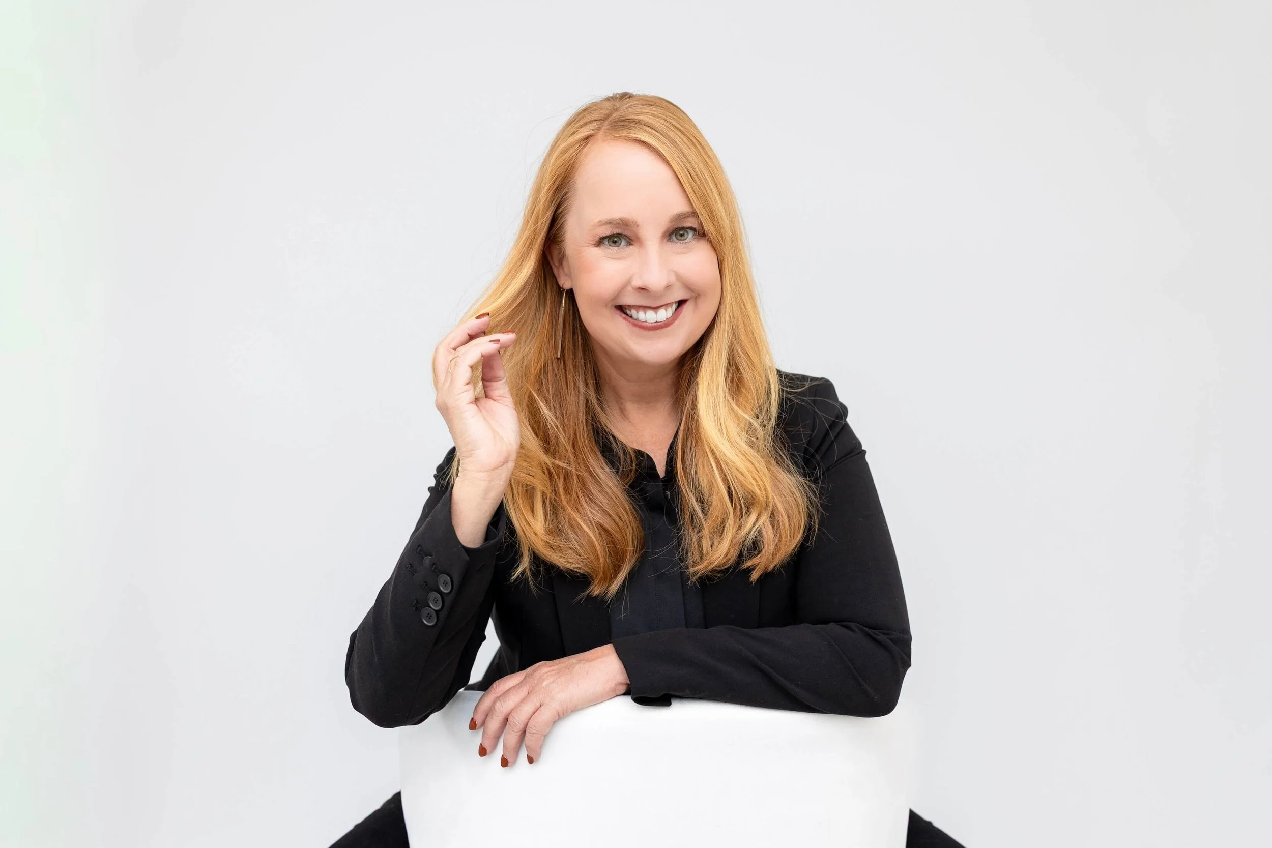 A red-haired woman wearing a black blazer, smiling, with her hand touching her hair, seated at a white table against a plain white background.