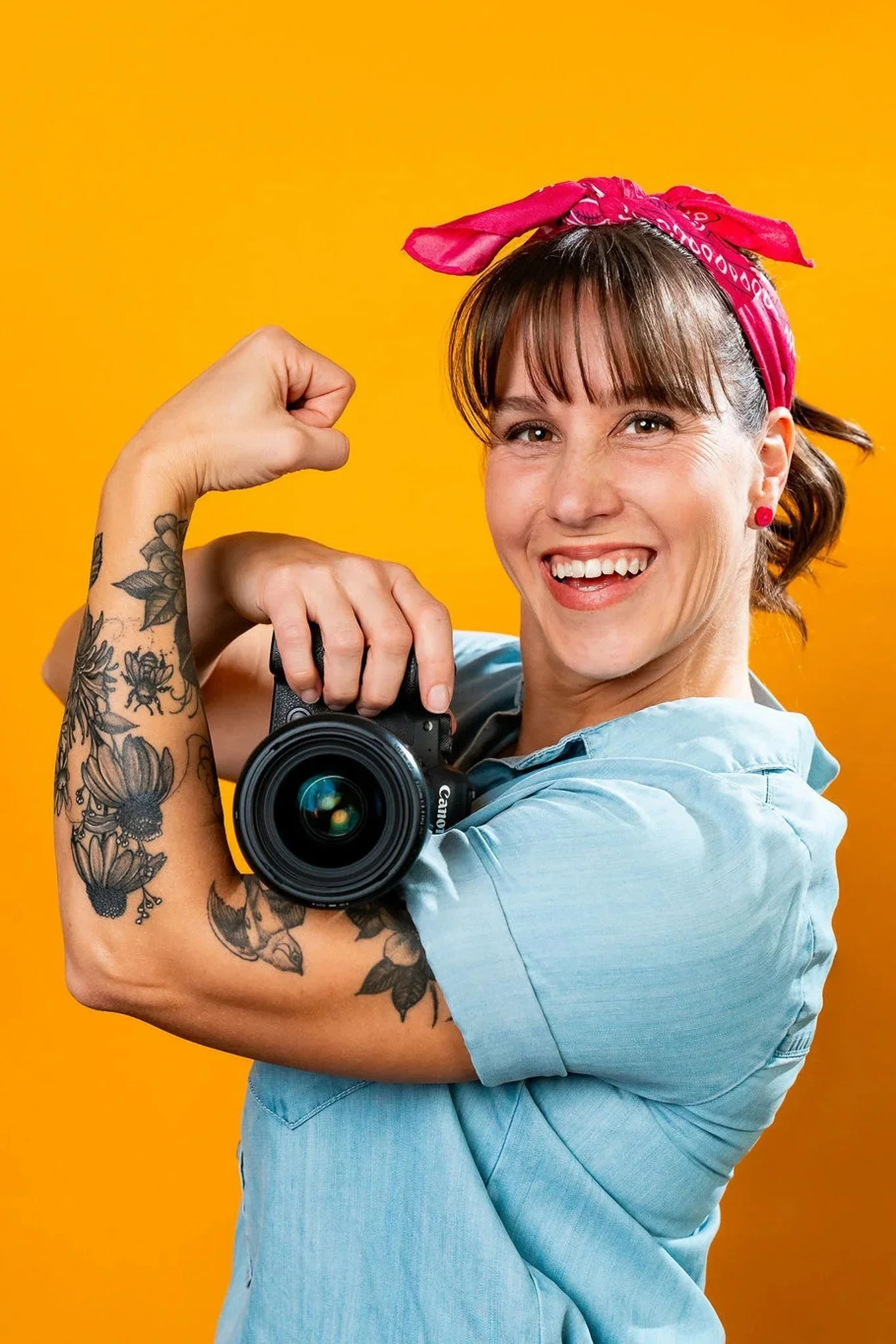 A woman with tattoos flexes her bicep while holding a camera, wearing a pink bandana and light blue shirt against a yellow background.