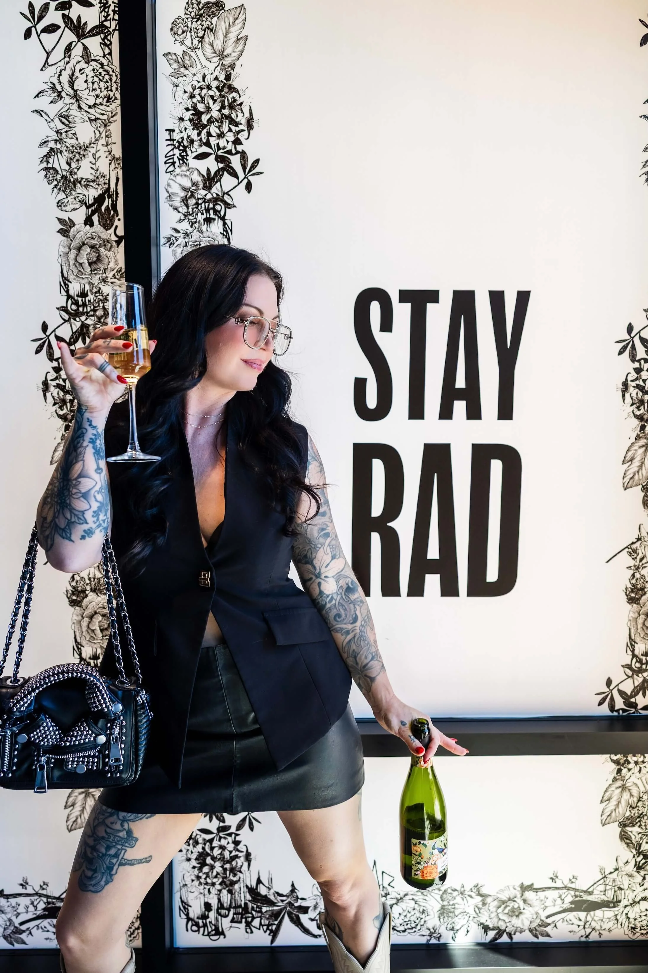 A woman with black hair, glasses, and tattoos holds a glass of champagne and a bottle of champagne, standing in front of a decorative floral wall with the words 'STAY BAD' in large black letters.