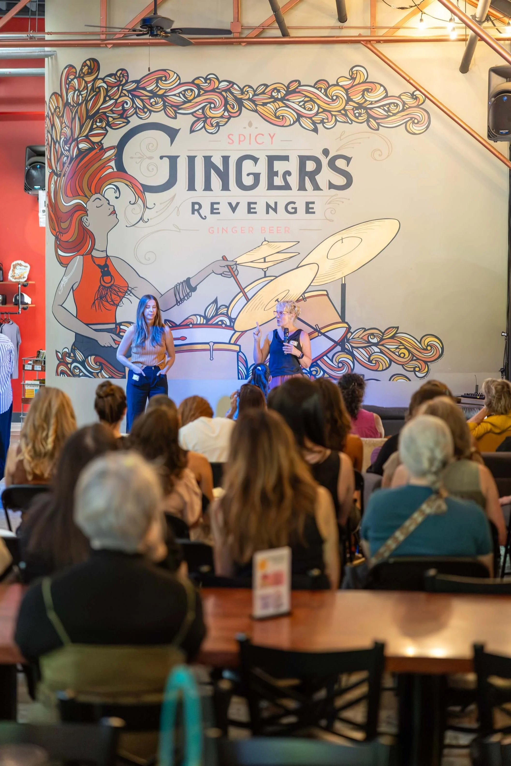 Large banner with artwork and text reading 'Spicy Ginger's Revenge Ginger Beer'. The artwork features a woman with long flowing red hair playing drums, surrounded by swirling designs. There are people sitting and standing in front of the banner, possibly at an event or presentation.