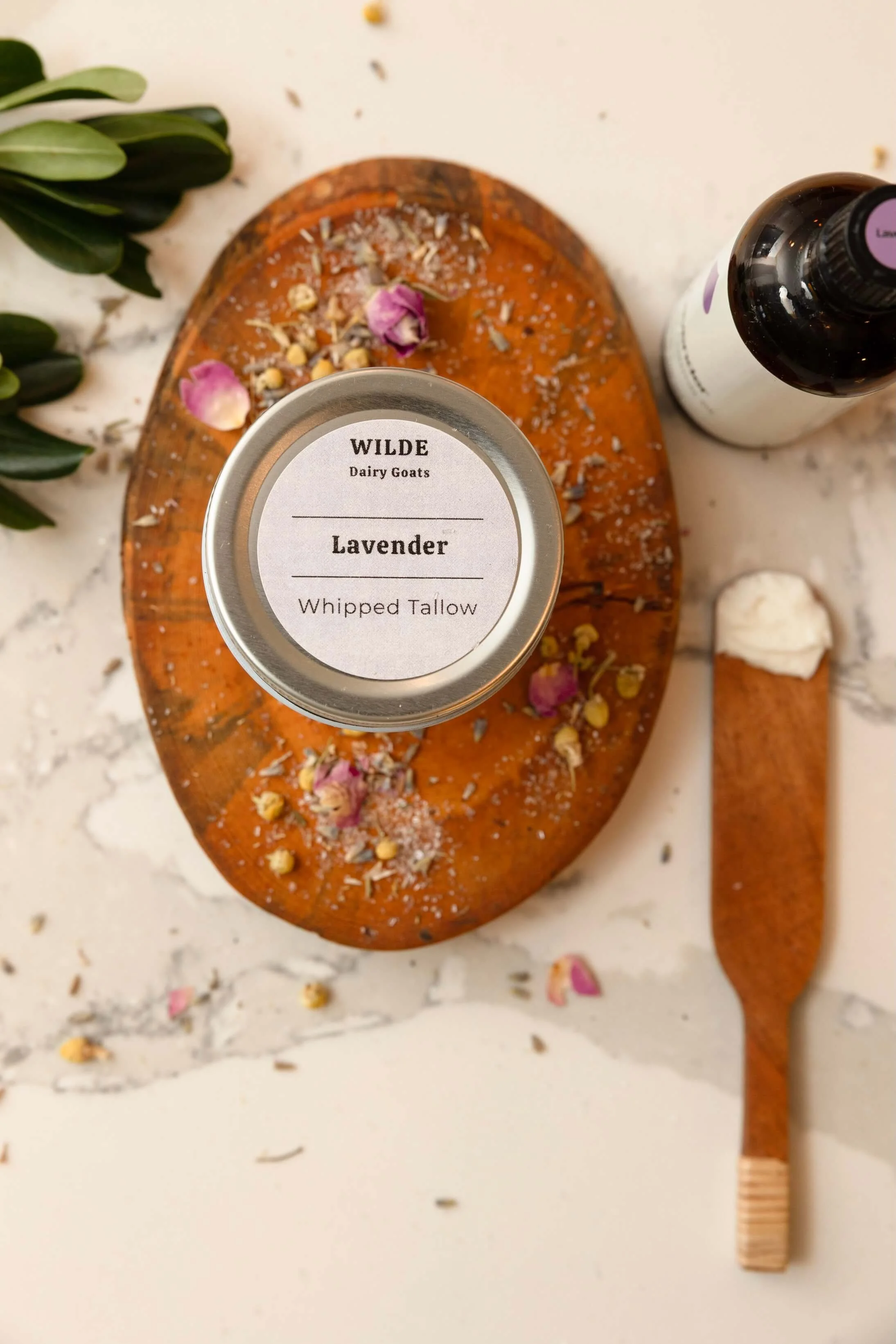 A jar labeled 'Wilde Dairy Goats Lavender Whipped Tallow' on a wooden cutting board with scattered flower petals, dried herbs, and a small wooden tool on a white marble surface.