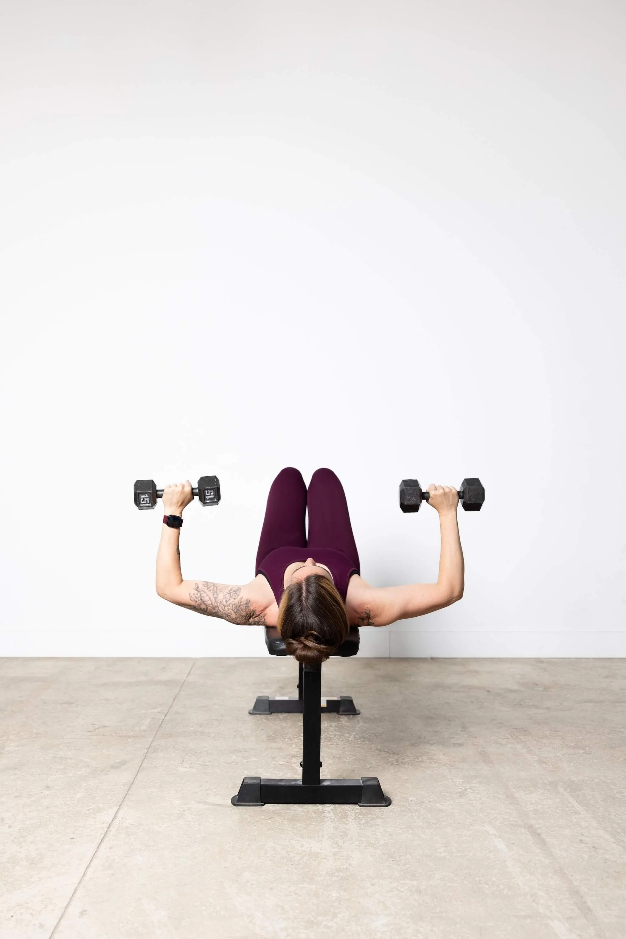A woman with tattoos on her arms lying on a workout bench, lifting dumbbells in each hand, with a blank white wall in the background and a concrete floor.