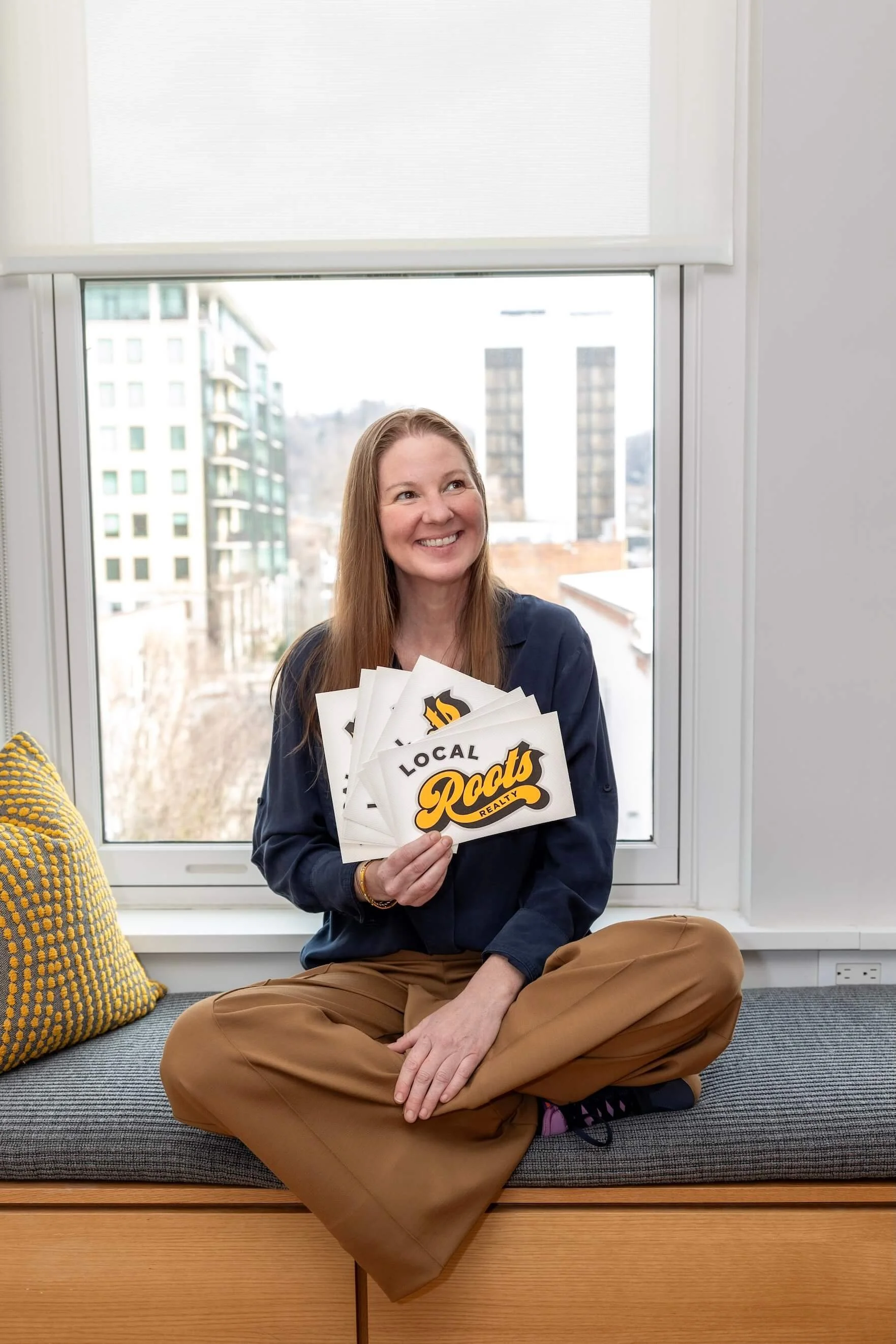 A smiling woman with long red hair, sitting cross-legged on a cushioned window seat, holding up flyers that say 'Local Roots Realty'. There is a large window behind her with a cityscape view of tall buildings.