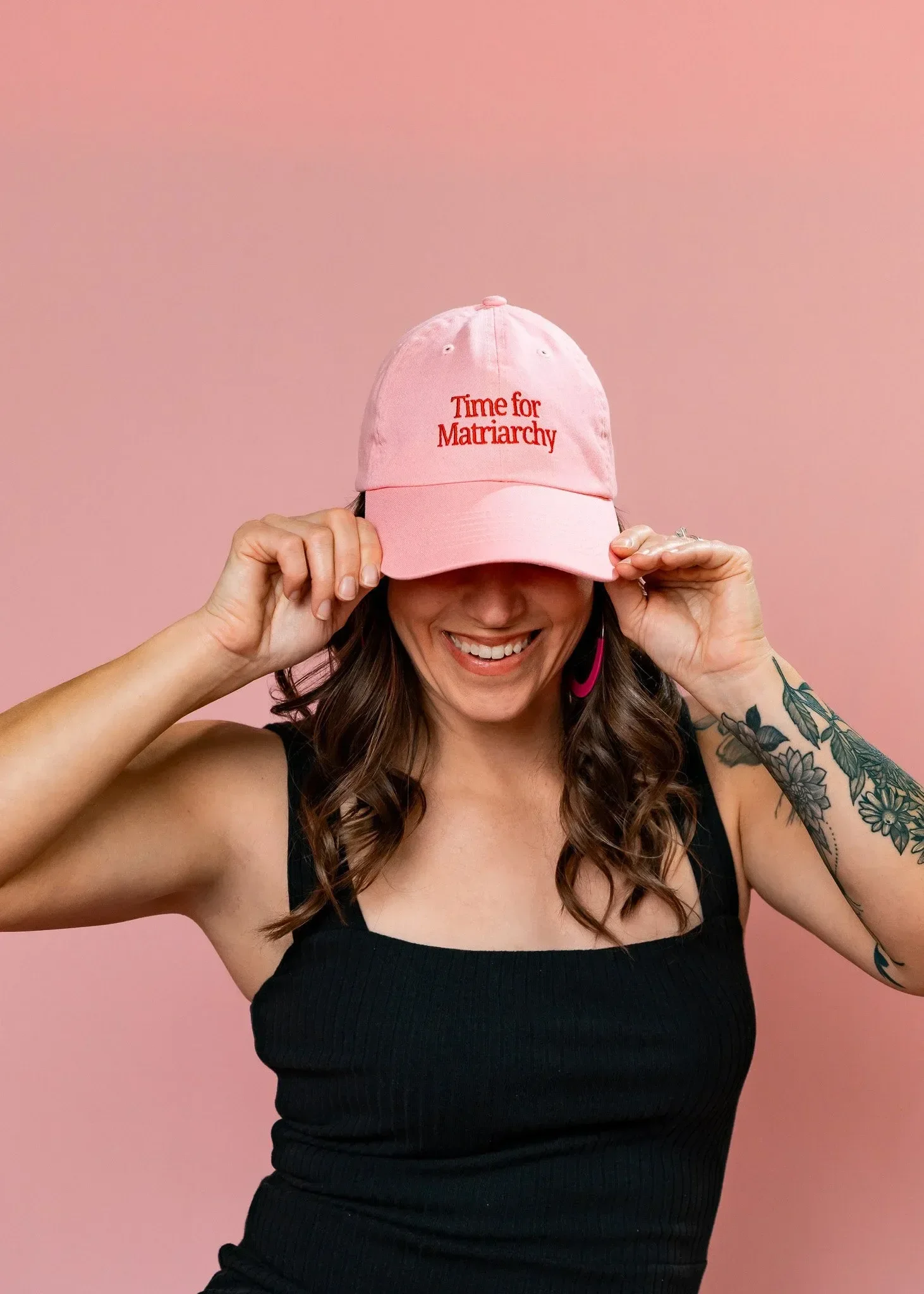 A woman smiling, wearing a pink baseball cap with red embroidered text that reads 'Time for Matriarchy,' black sleeveless top, holding the brim of her cap, against a pink background, with floral tattoos on her left arm and pink earrings.