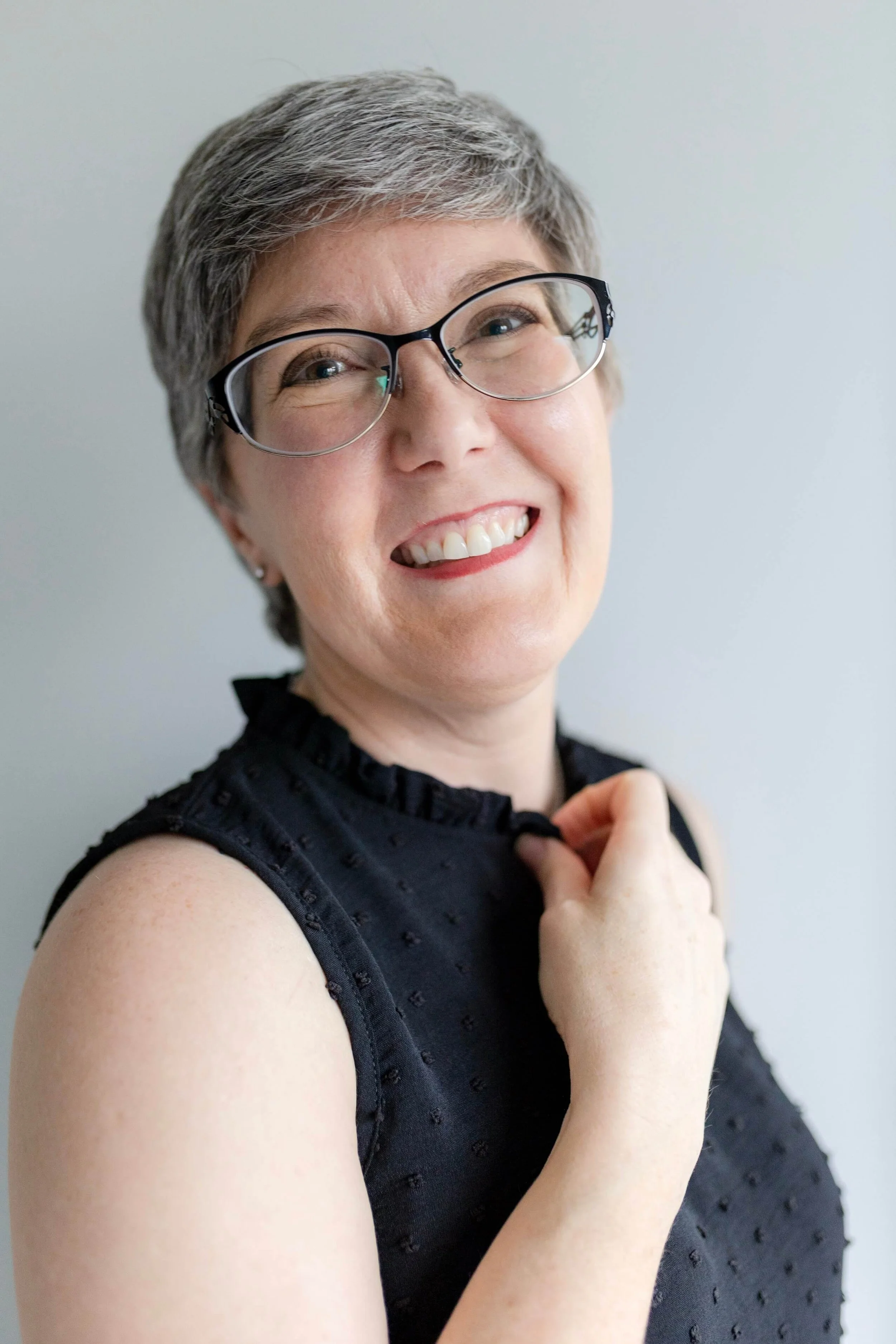 Close-up of a smiling woman with short gray hair, glasses, and fair skin, wearing a sleeveless black dress, standing against a plain light background.