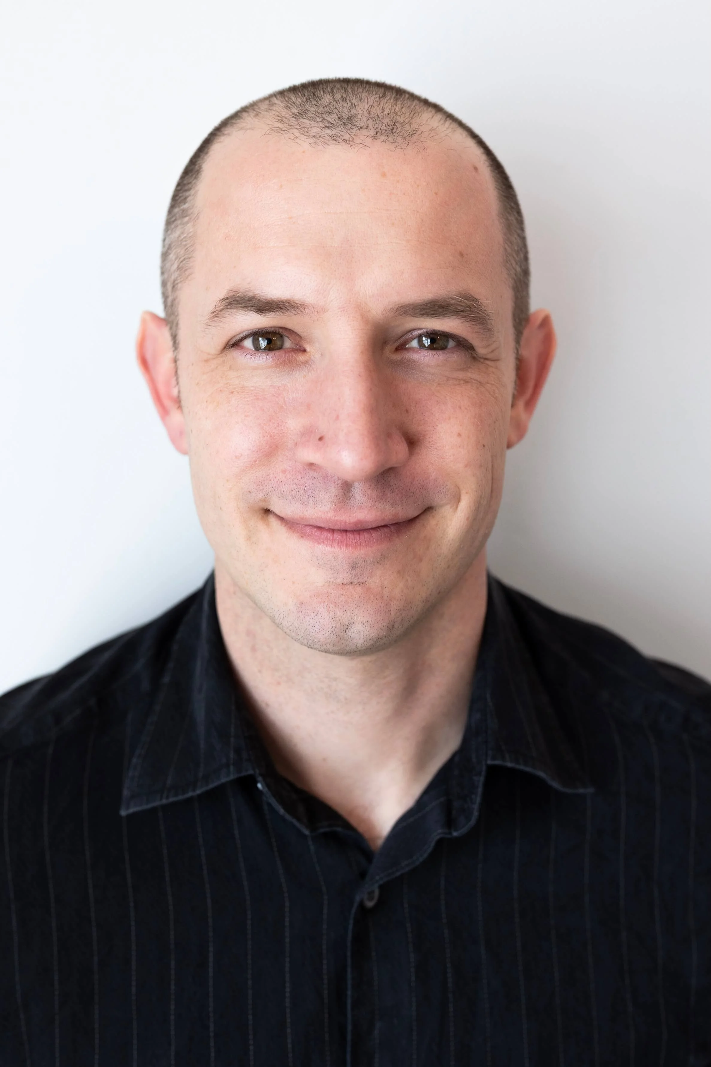Close-up photo of a smiling man with a shaved head, wearing a dark striped shirt, standing against a plain white background.