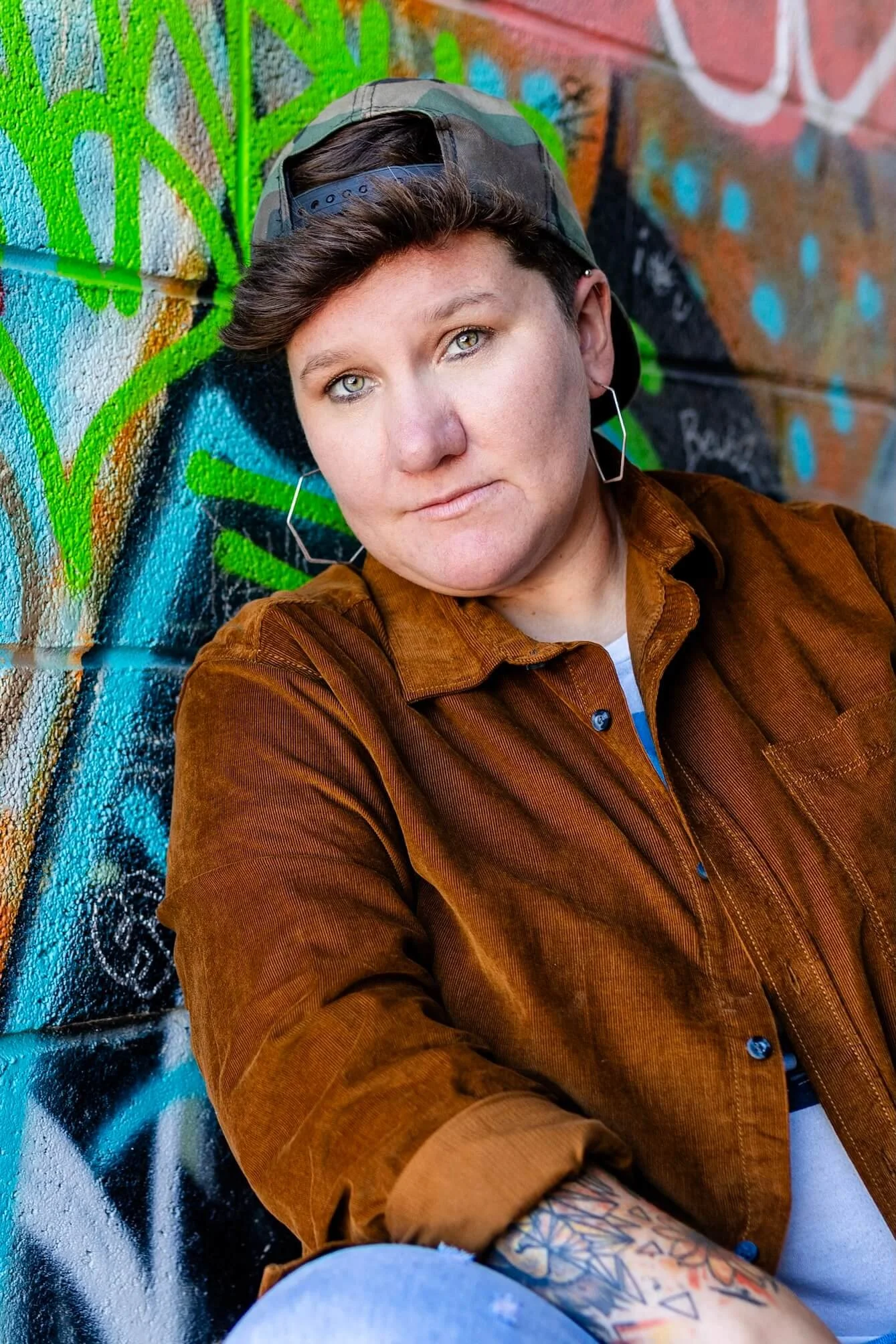A person with short hair and light eyes taking a selfie against a colorful graffiti wall, wearing a brown shirt, cap worn backwards, and earrings.