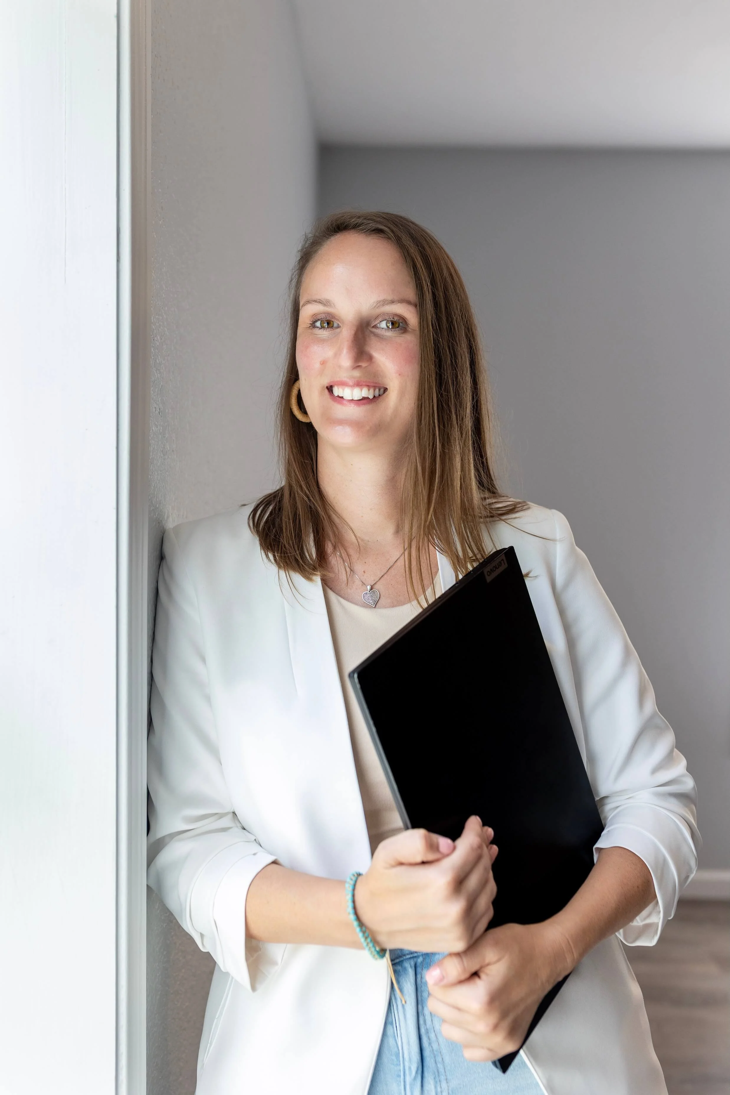 A woman with shoulder-length brown hair, wearing a white blazer, holding a black folder, standing against a gray wall, smiling at the camera.