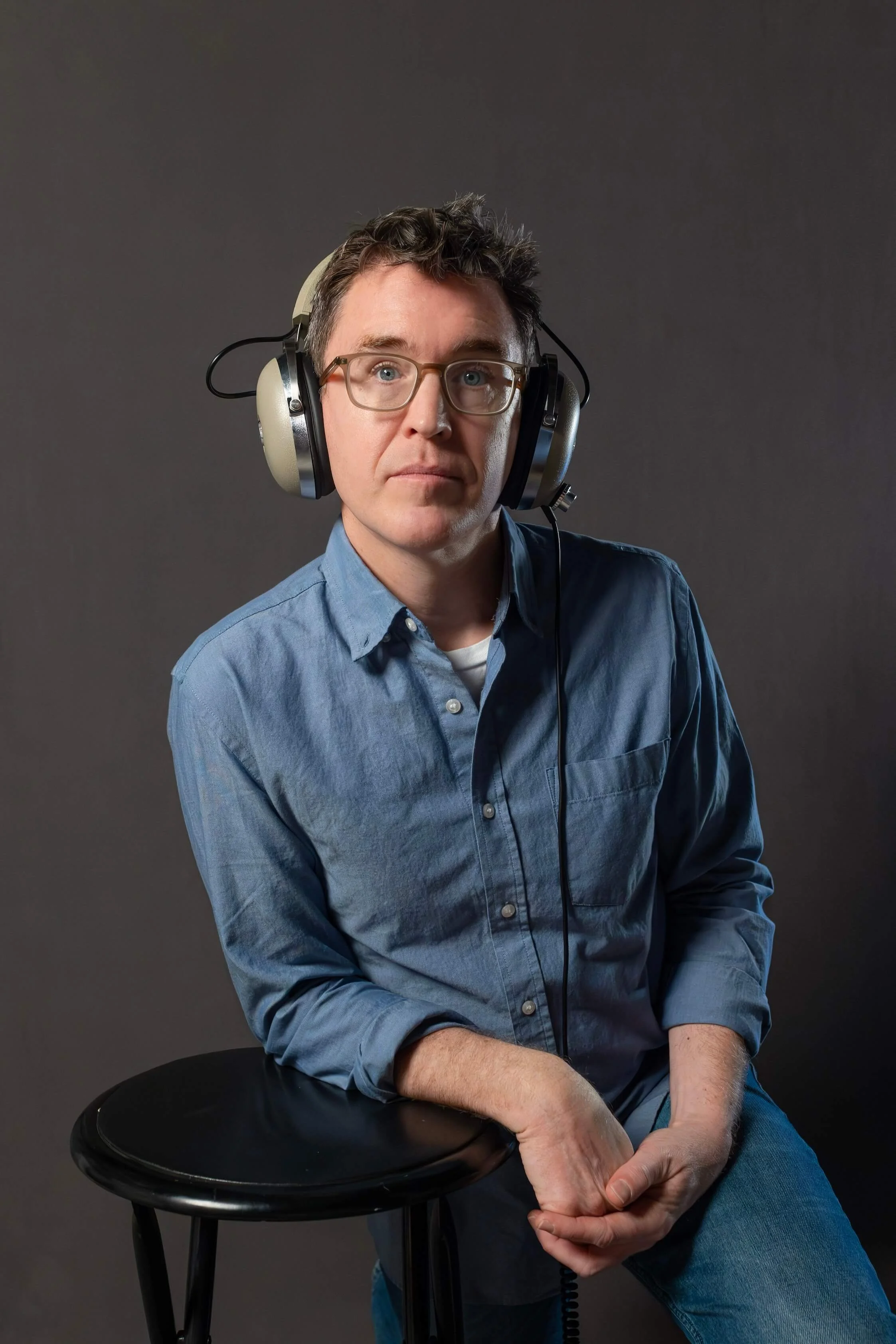 A man wearing glasses and headphones sits on a stool with a dark gray background.