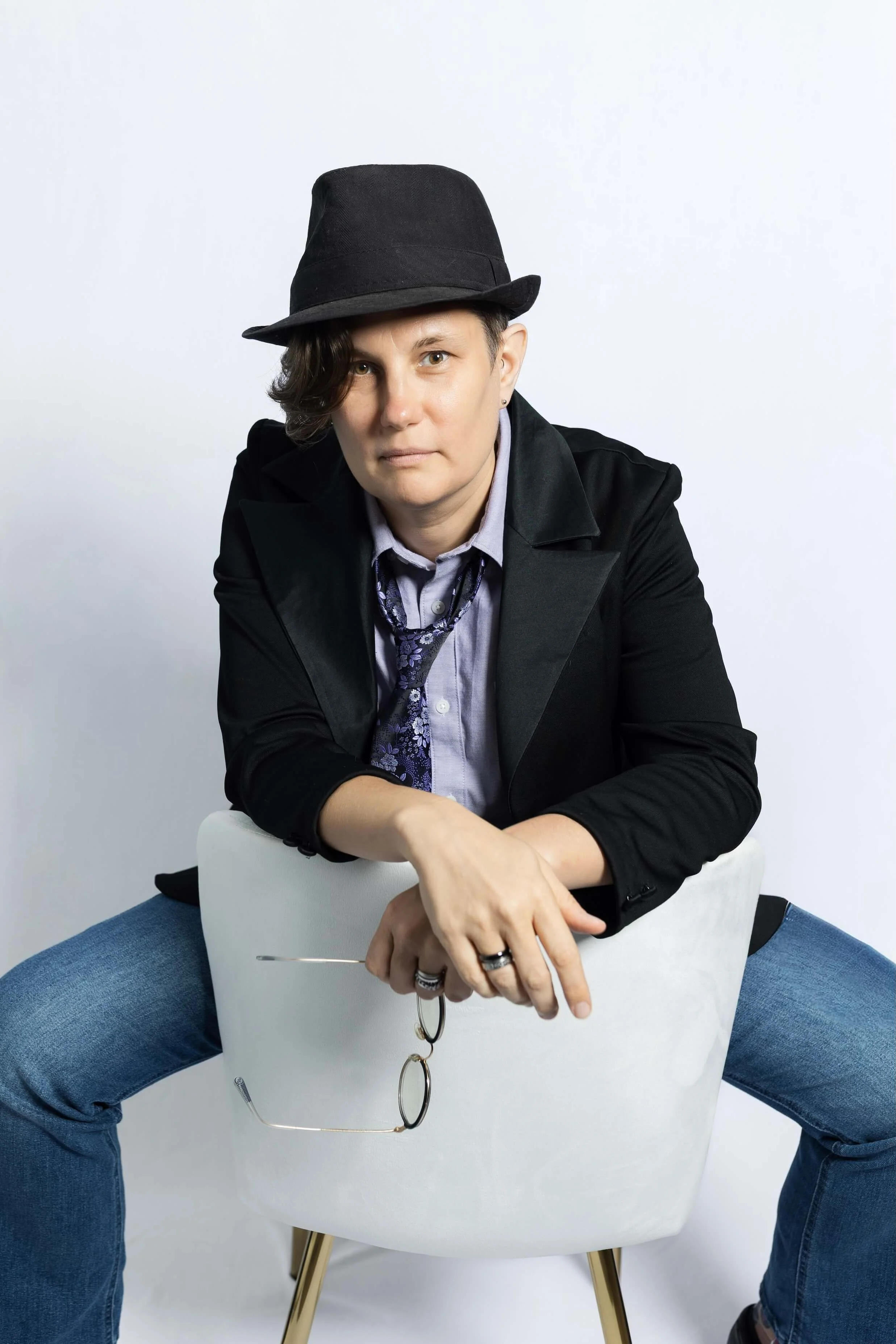 Person sitting backwards on a white chair, wearing a black hat, black jacket, light shirt, and blue jeans. The person's arms rest on the chair, holding eyeglasses and keys, with a white background.