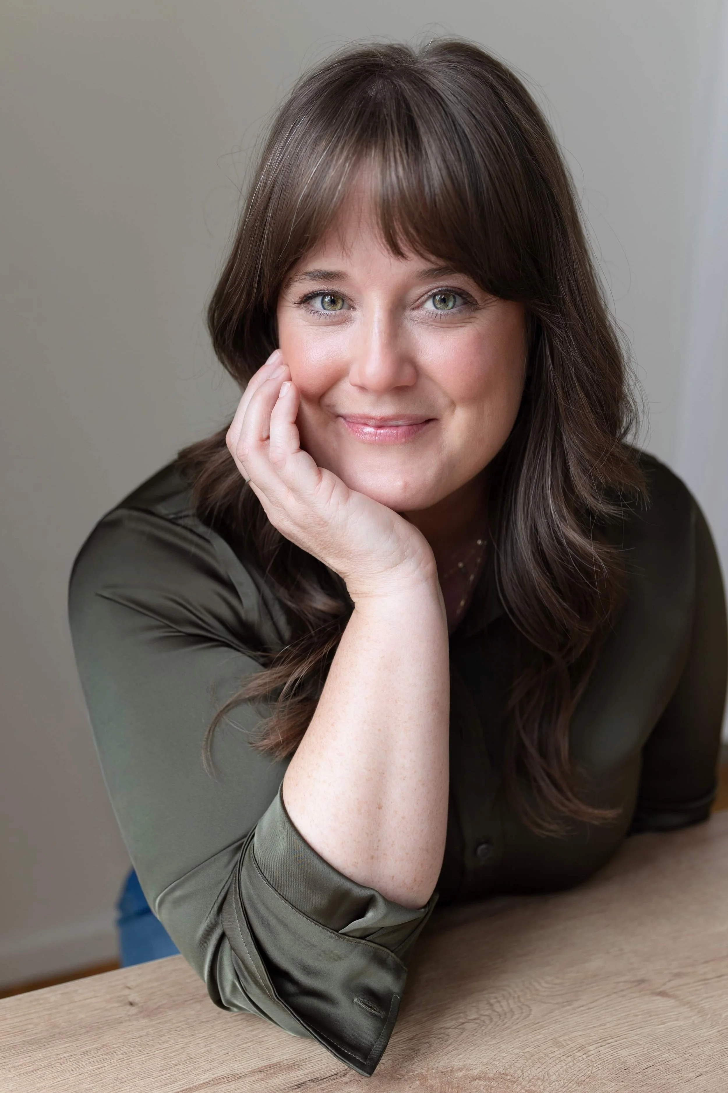 A woman with green eyes and long brown hair, smiling and resting her head on her hand, sitting at a table.