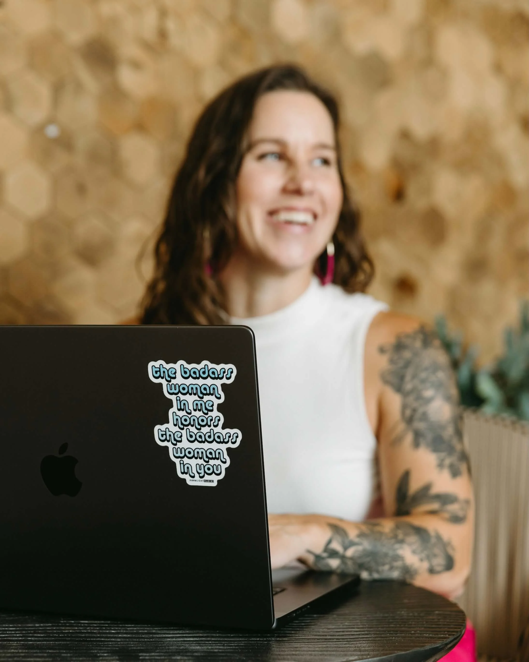 A woman with dark hair and tattoos on her arm smiling at a laptop that has a sticker reading 'the badass woman in me honors the badass woman in you' in a textured wooden background.