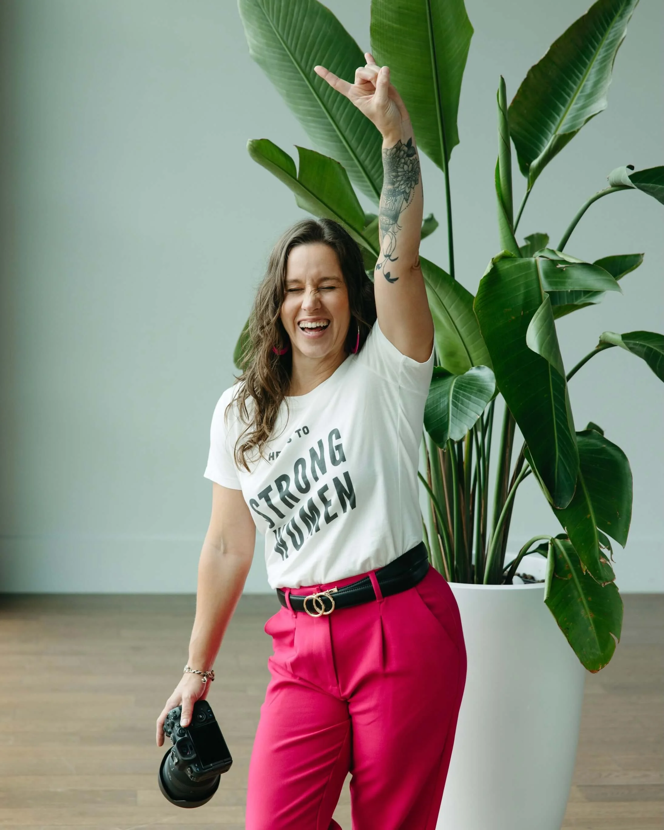 Woman with long hair and tattoos on her arm, dressed in a white shirt with text, bright pink pants, holding a camera, standing next to a large potted plant with big green leaves. She has a big smile, eyes closed, and is making a rock-and-roll hand gesture with her other hand.