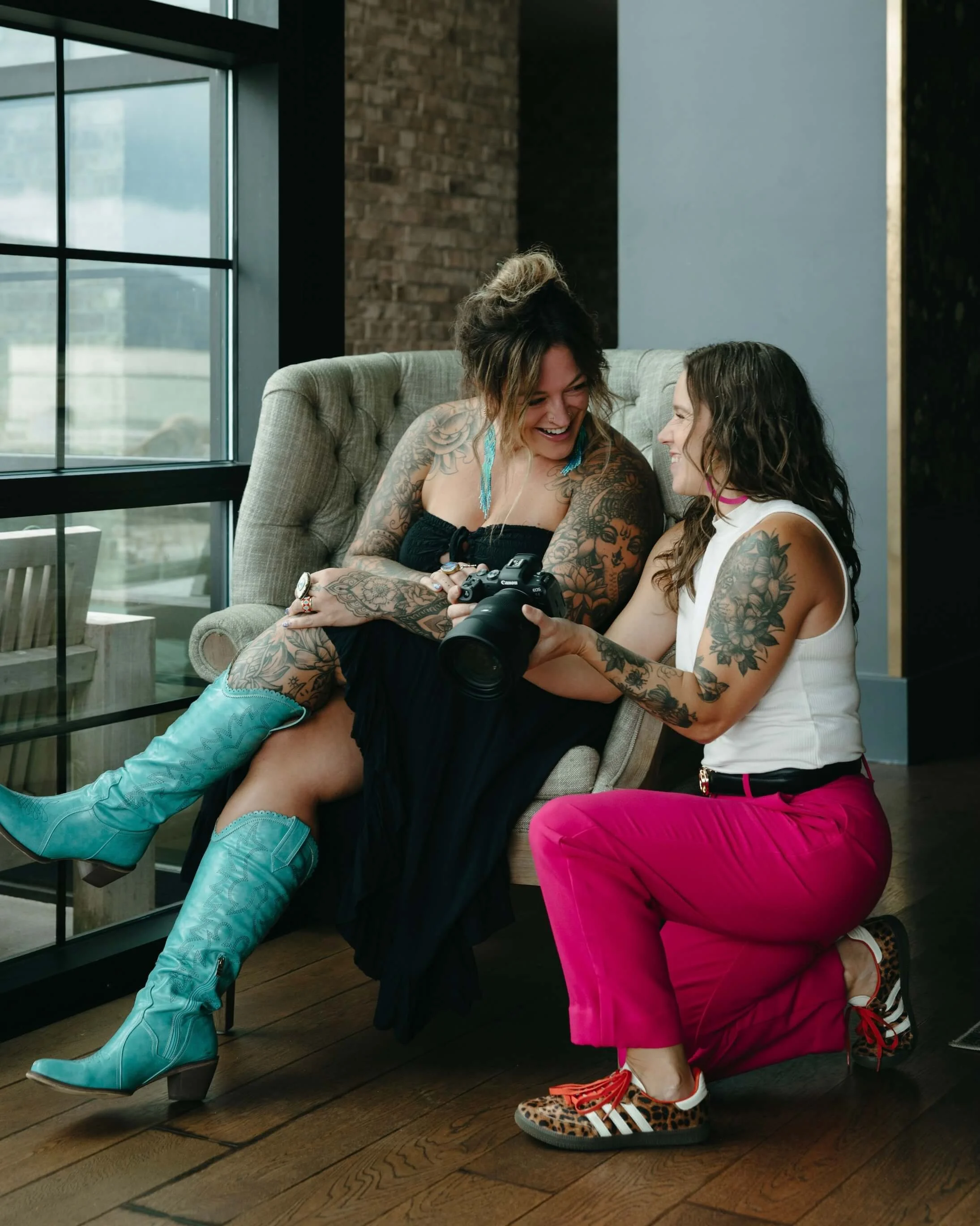 Two women with tattoos having fun and sharing a camera in a modern indoor setting with large windows, a brick wall, and wooden floors.