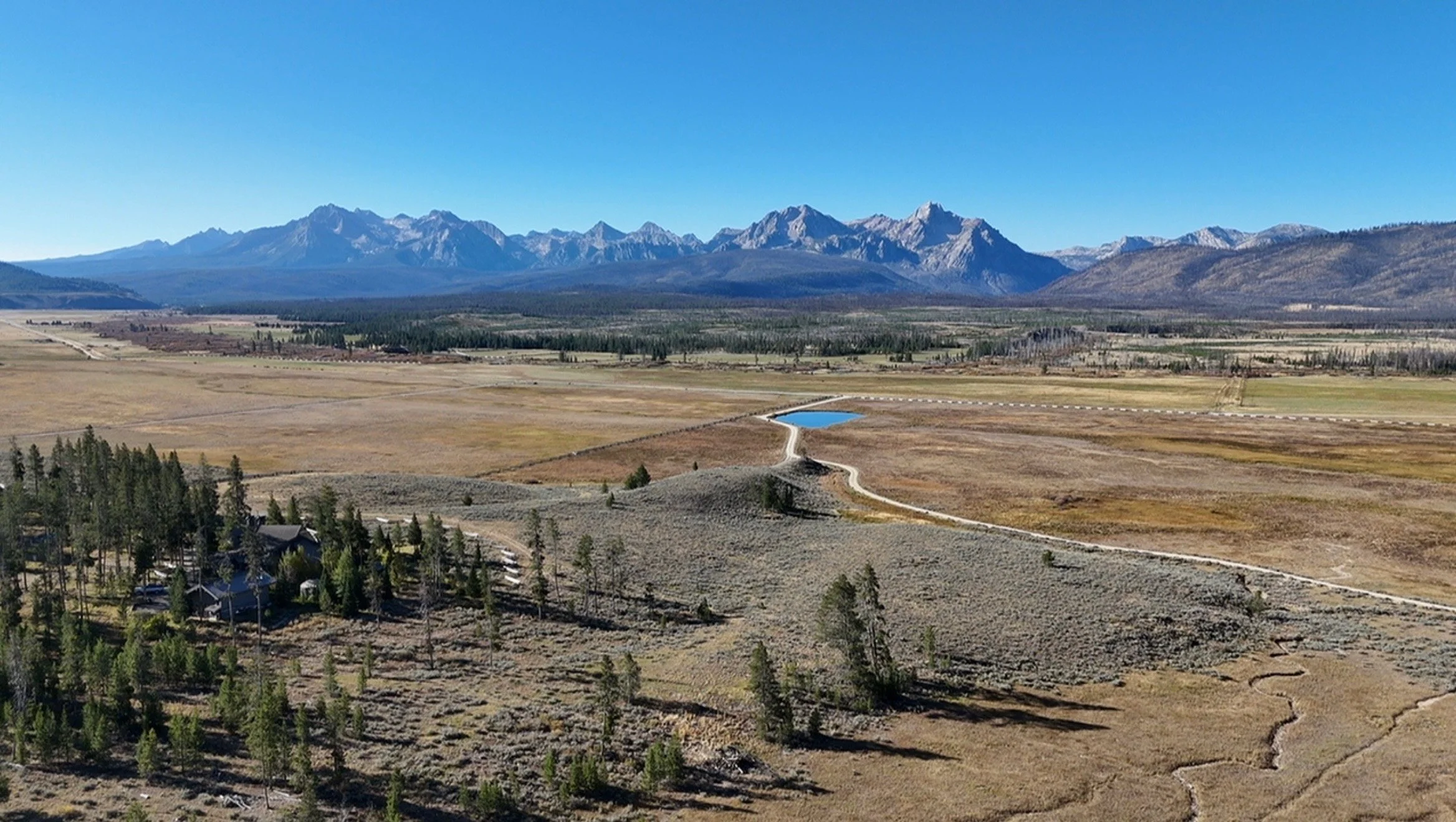 A landscape with mountains in the background, a flat valley in the middle, and a small road with a pond in the foreground. There are some trees and buildings in the lower part of the image.