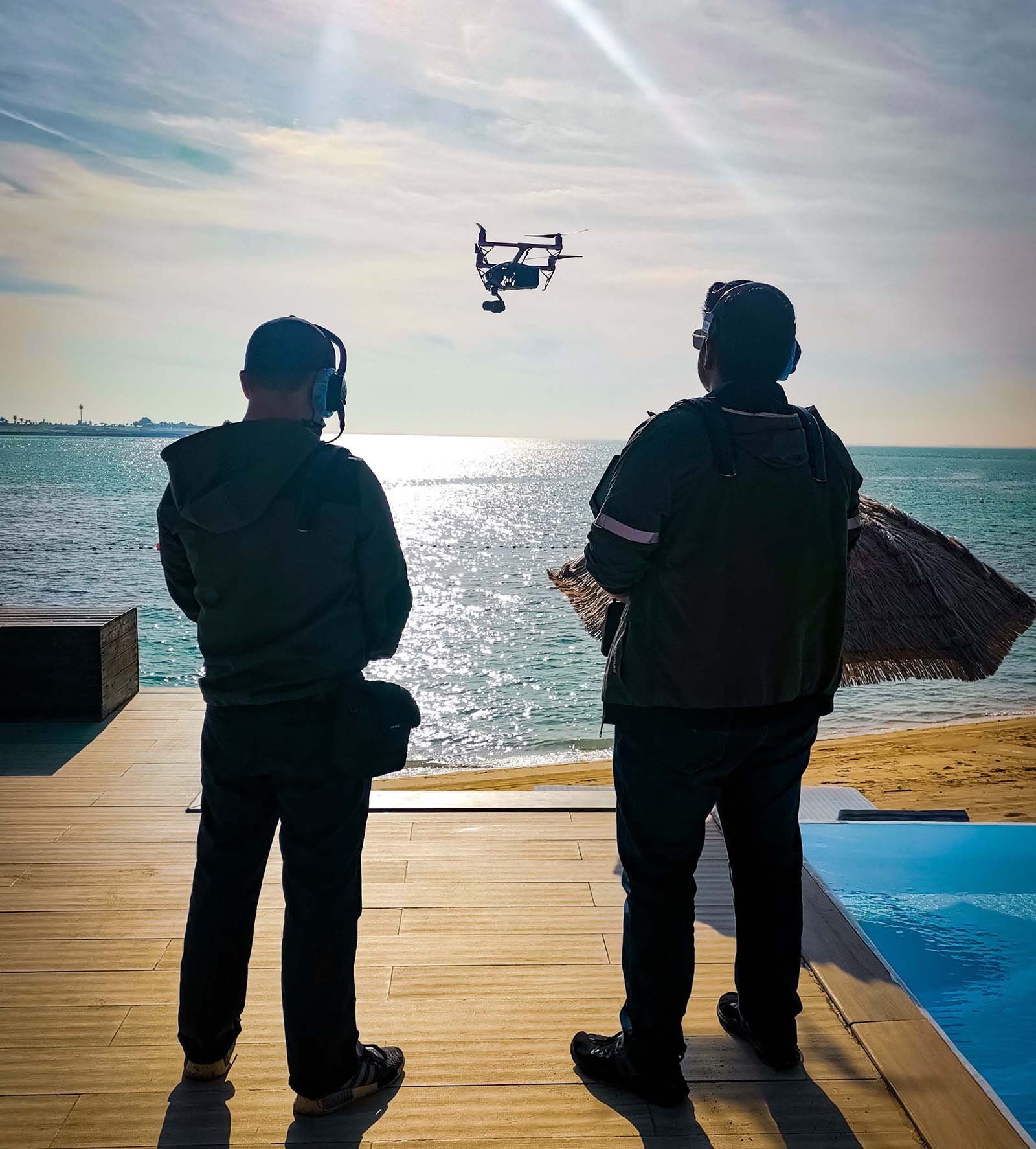  Two drone operators with control panels overseeing a shoot on the beach, coordinating aerial filming operations. 