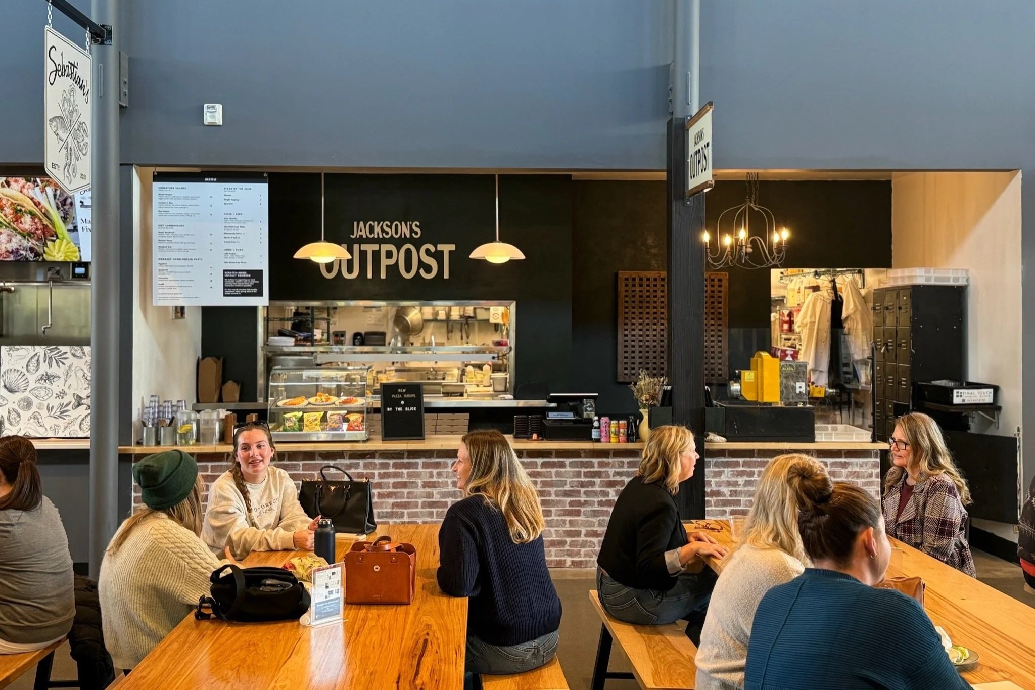 Indoor seating area at Jackson's Outpost with several women sitting at wooden tables, talking. The background features a black wall with the sign 'Jackson's Outpost', a menu display, and a counter with food and kitchen items.