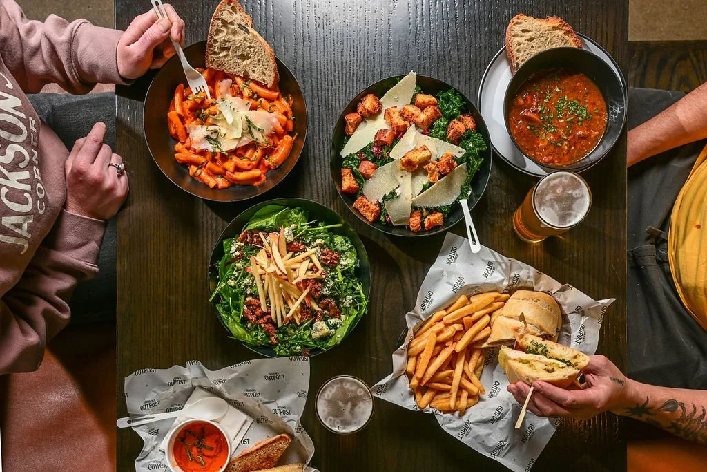 A table with a variety of dishes including pasta with tomato sauce, Caesar salad, soup in a black bowl with bread, a sandwich with fries, and a green salad with cheese and nuts.