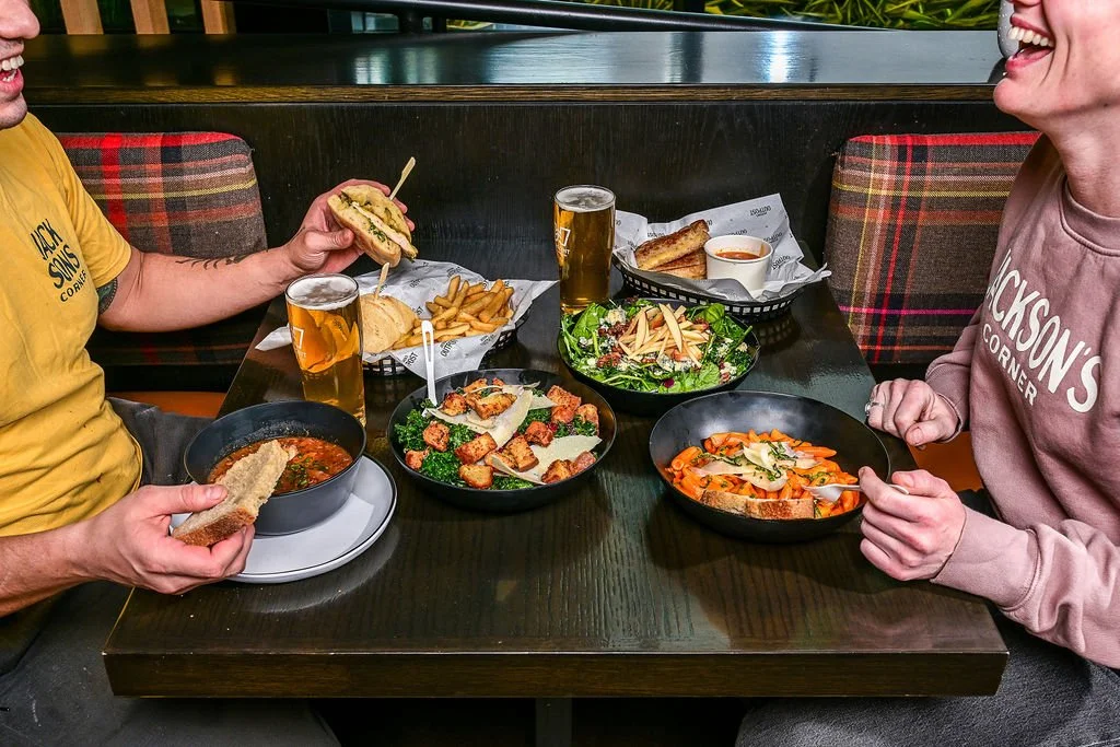 Two people dining together at a restaurant, with various dishes including soup, sandwiches, salad, pasta, and drinks on the table.