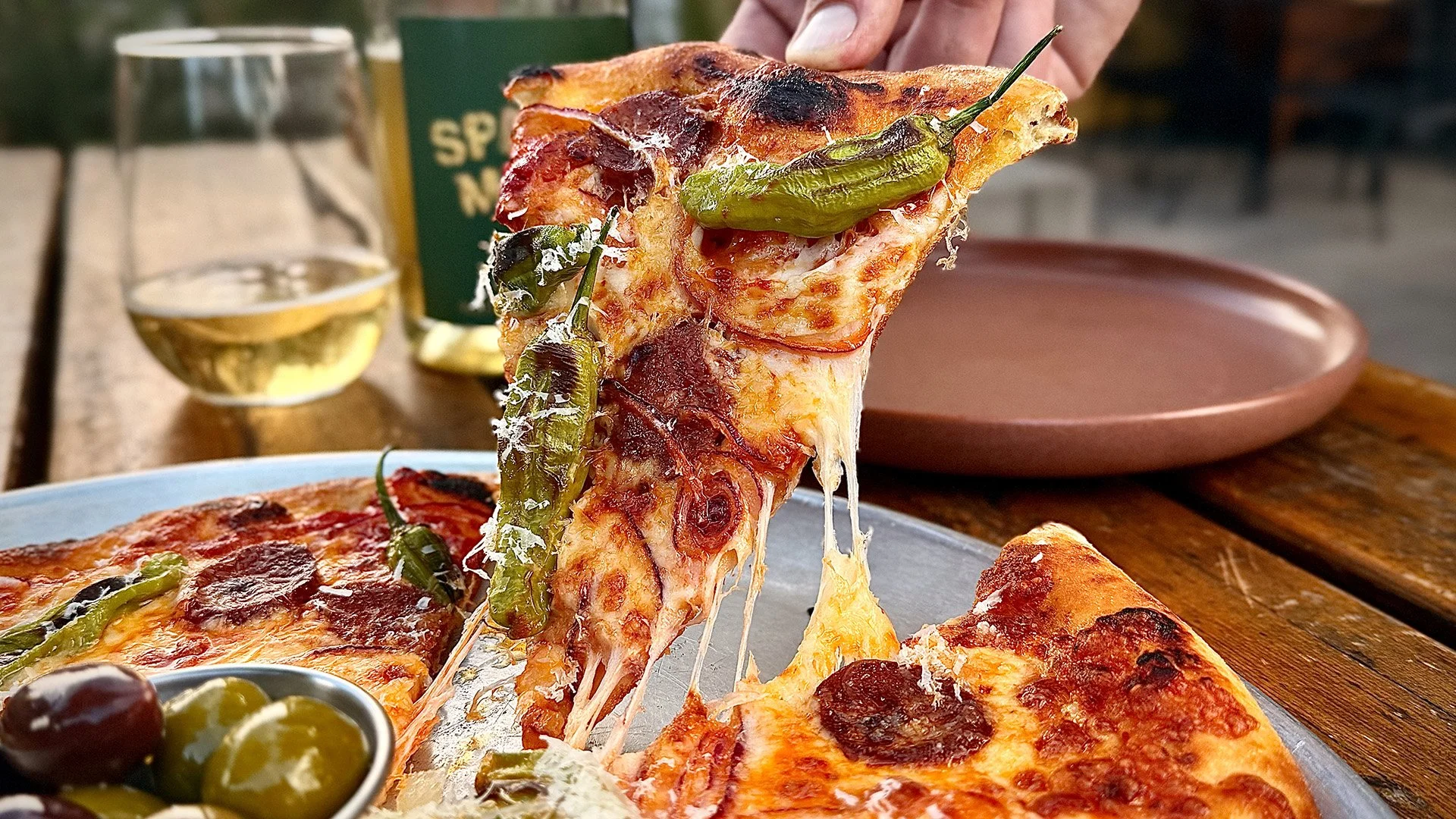 Close-up of a slice of pizza with green peppers, pepperoni, and melted cheese, being lifted from a pizza platter outdoors on a wooden table, accompanied by a glass of white wine, a bowl of mixed olives, and an empty pink plate.