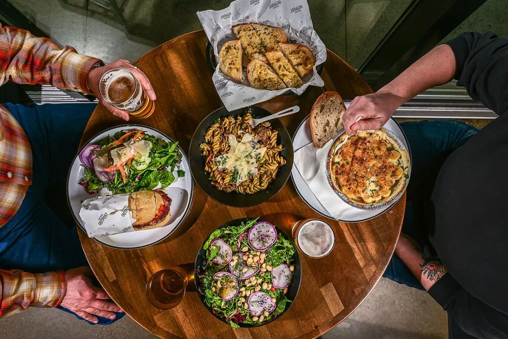 A round wooden table with two people dining. The table has a salad with greens, radishes, and onions, a slice of grilled vegetable pizza, a bowl of pasta with cheese, a basket of sliced bread, a mixed green salad with radishes and chickpeas, and a sl