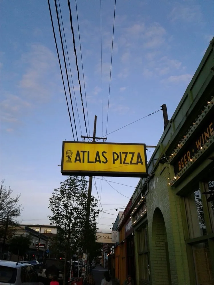 Yellow sign for Atlas Pizza hanging above a city street with trees and buildings, with a clear blue sky overhead.