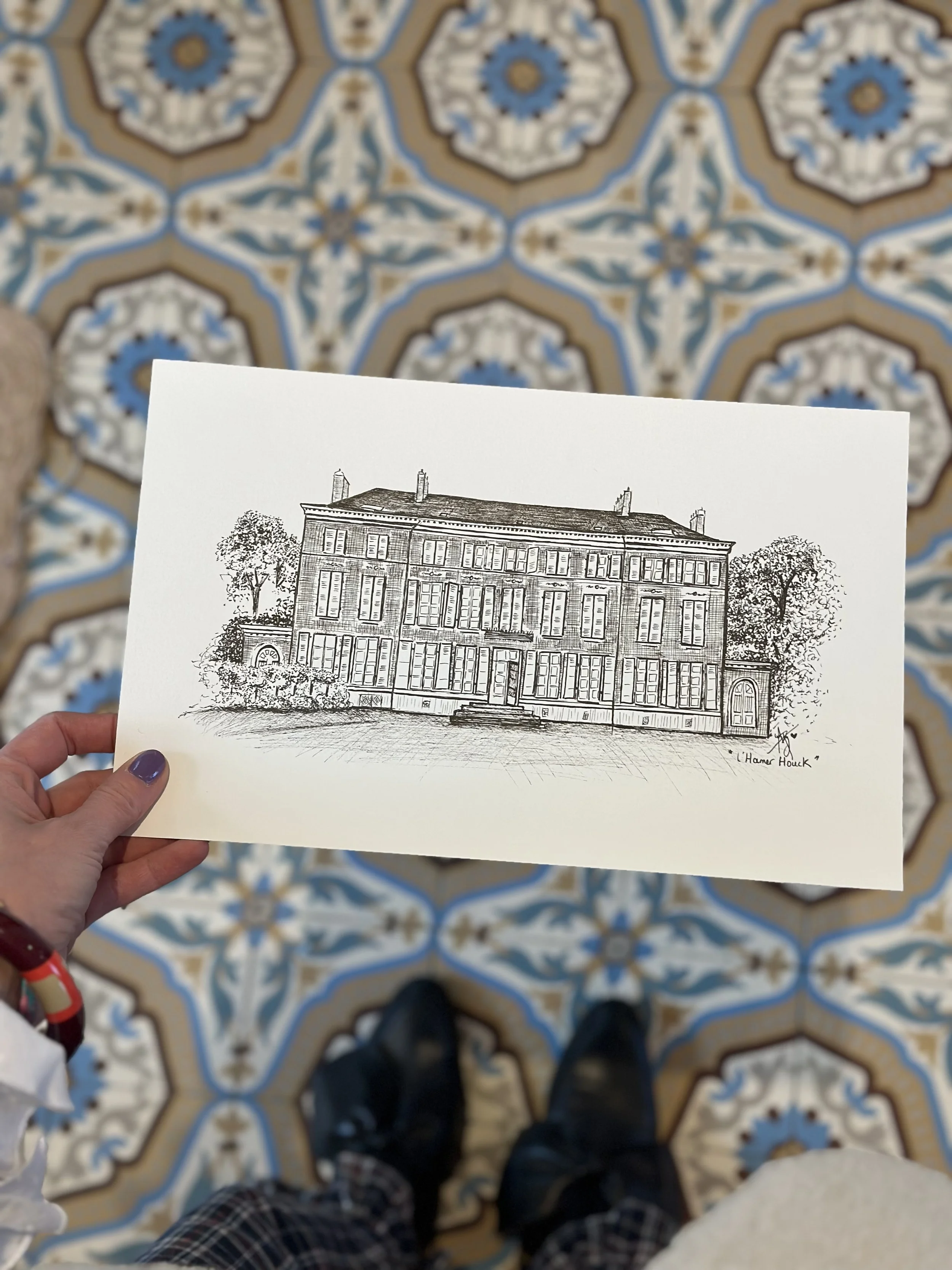 Une main tient une feuille avec un dessin d'un bâtiment, vu du dessus, avec deux arbres de chaque côté, devant un sol en pavés. Le bâtiment ressemble à un ancien hôtel ou palais.