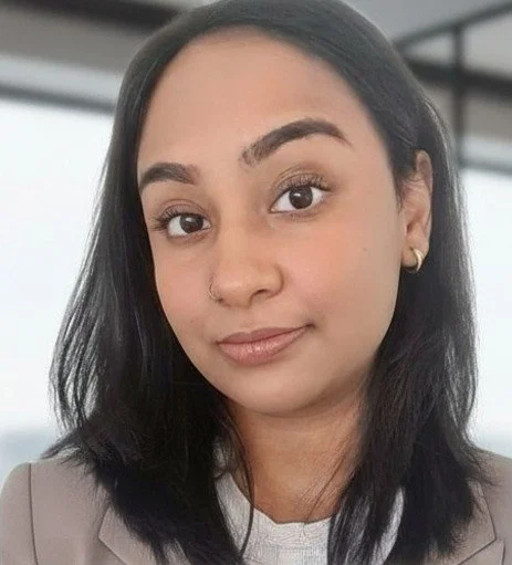 Close-up of a young woman with dark hair, light makeup, and wearing a blazer and small earrings.