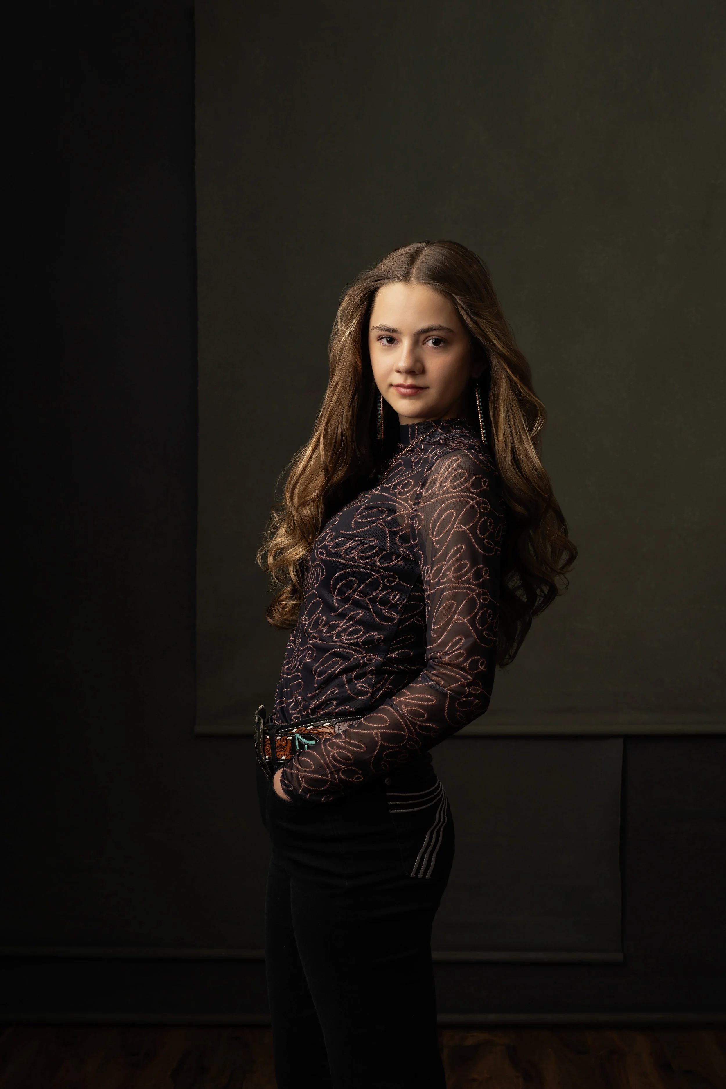 Young woman with long wavy brown hair, wearing a fitted black shirt with swirling light-colored cursive text, black pants with white stripes, and long earrings, standing against a dark background with a faint brown floor visible.