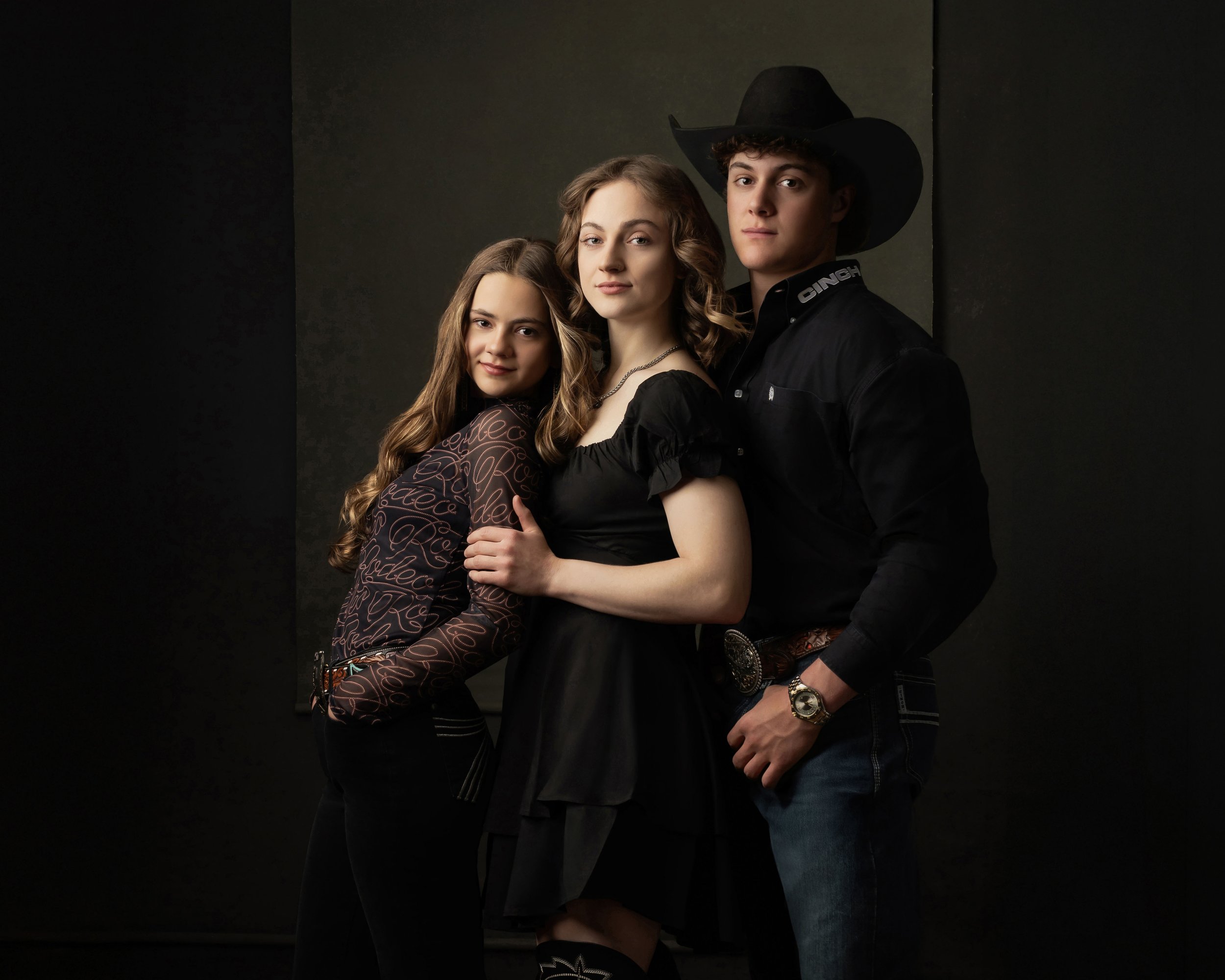 Three young adults posing together against a dark background, with two women and one man dressed in stylish clothing, the man wearing a large black cowboy hat.