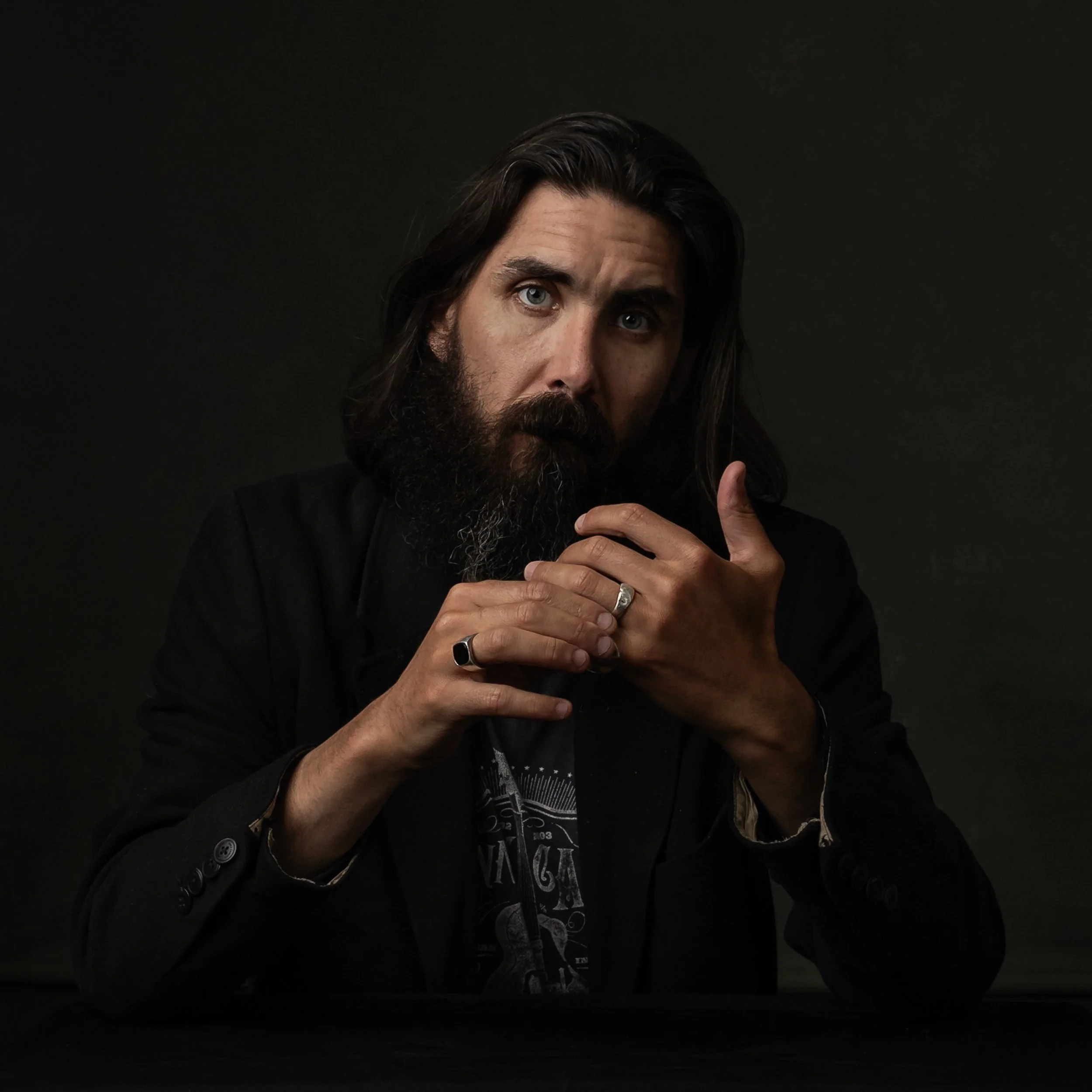 A man with a beard and long dark hair, wearing rings, a black jacket, and a graphic T-shirt, posing against a black background.