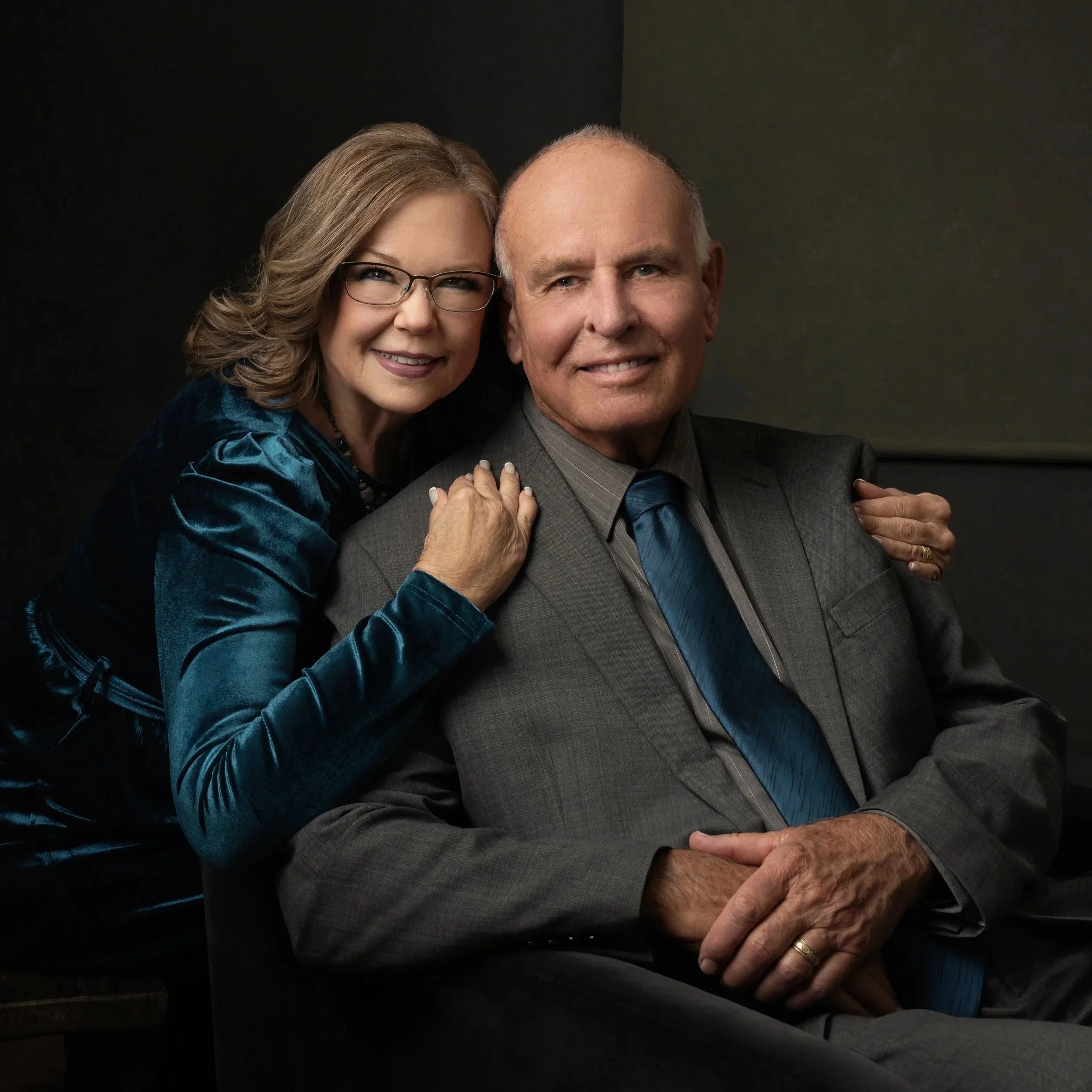 A woman with curly blonde hair, glasses, and a blue velvet dress is smiling and holding hands with a man with short gray hair, wearing a gray suit and tie, against a dark background.