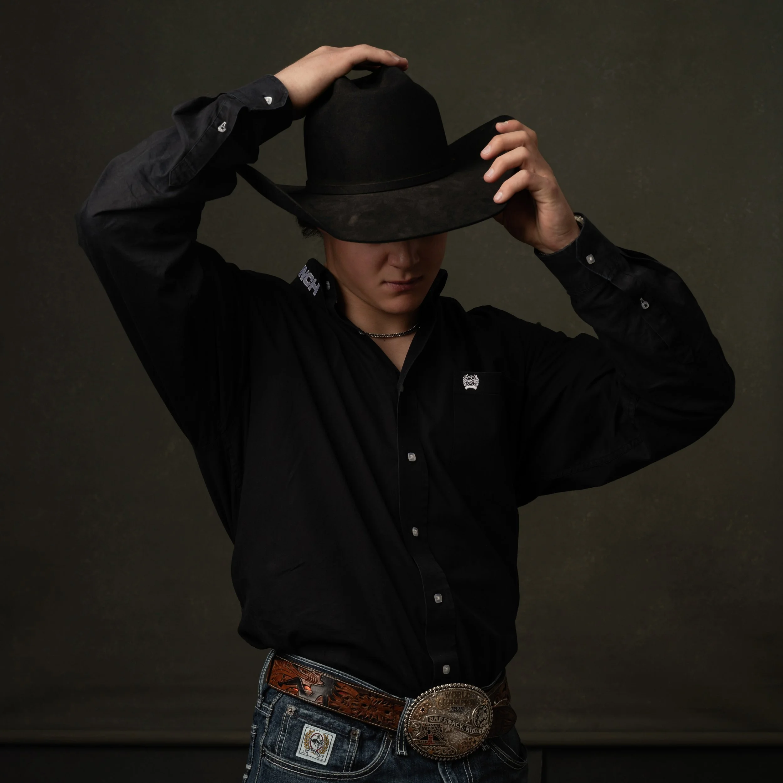A person wearing a black button-up shirt, holding a black cowboy hat on their head with both hands, and wearing a large rodeo champion belt with multiple medals and patches over their jeans. The background is plain and dark.