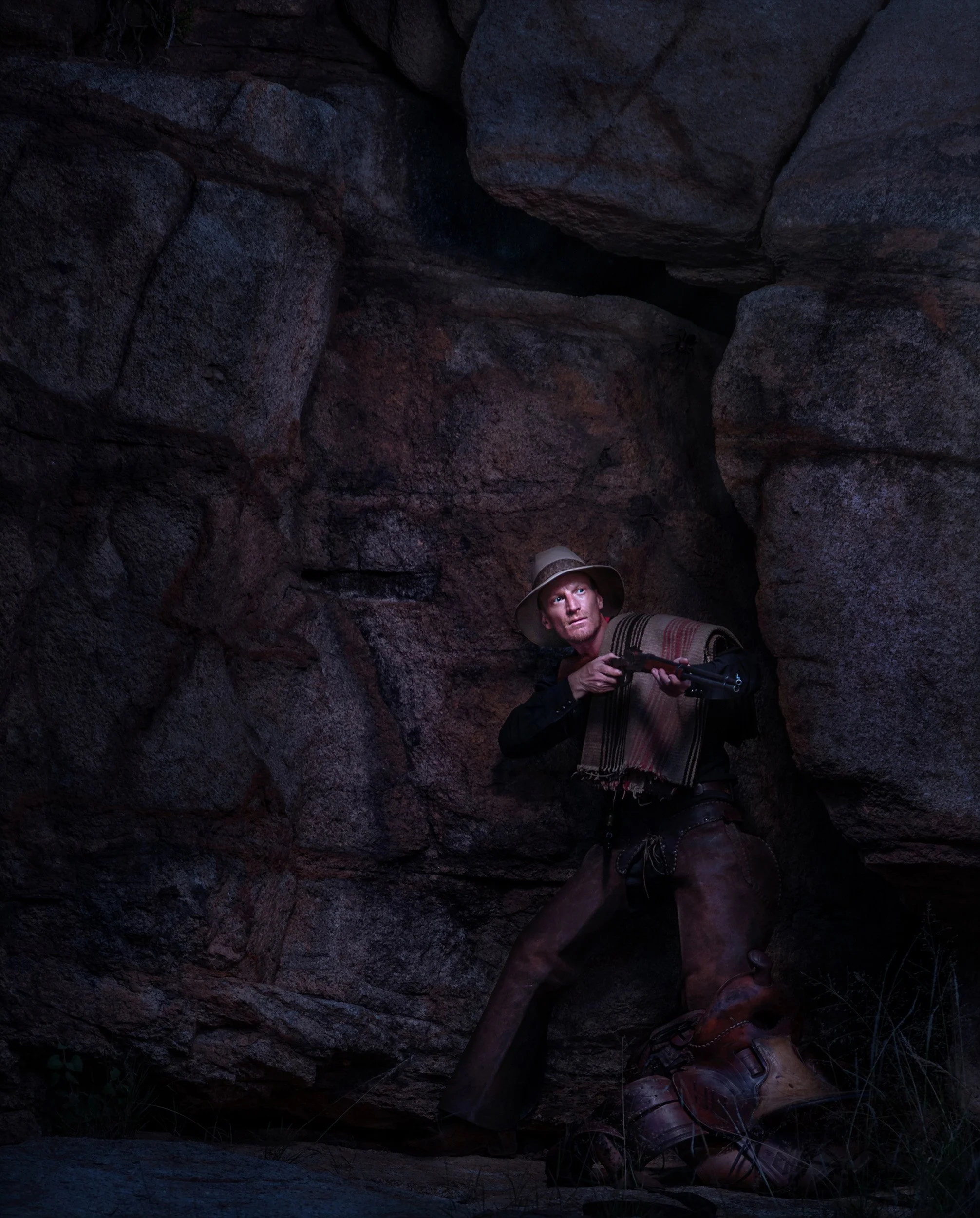 Cowboy with gun and saddle backed against a rock wall with tarantula coming down rock face
