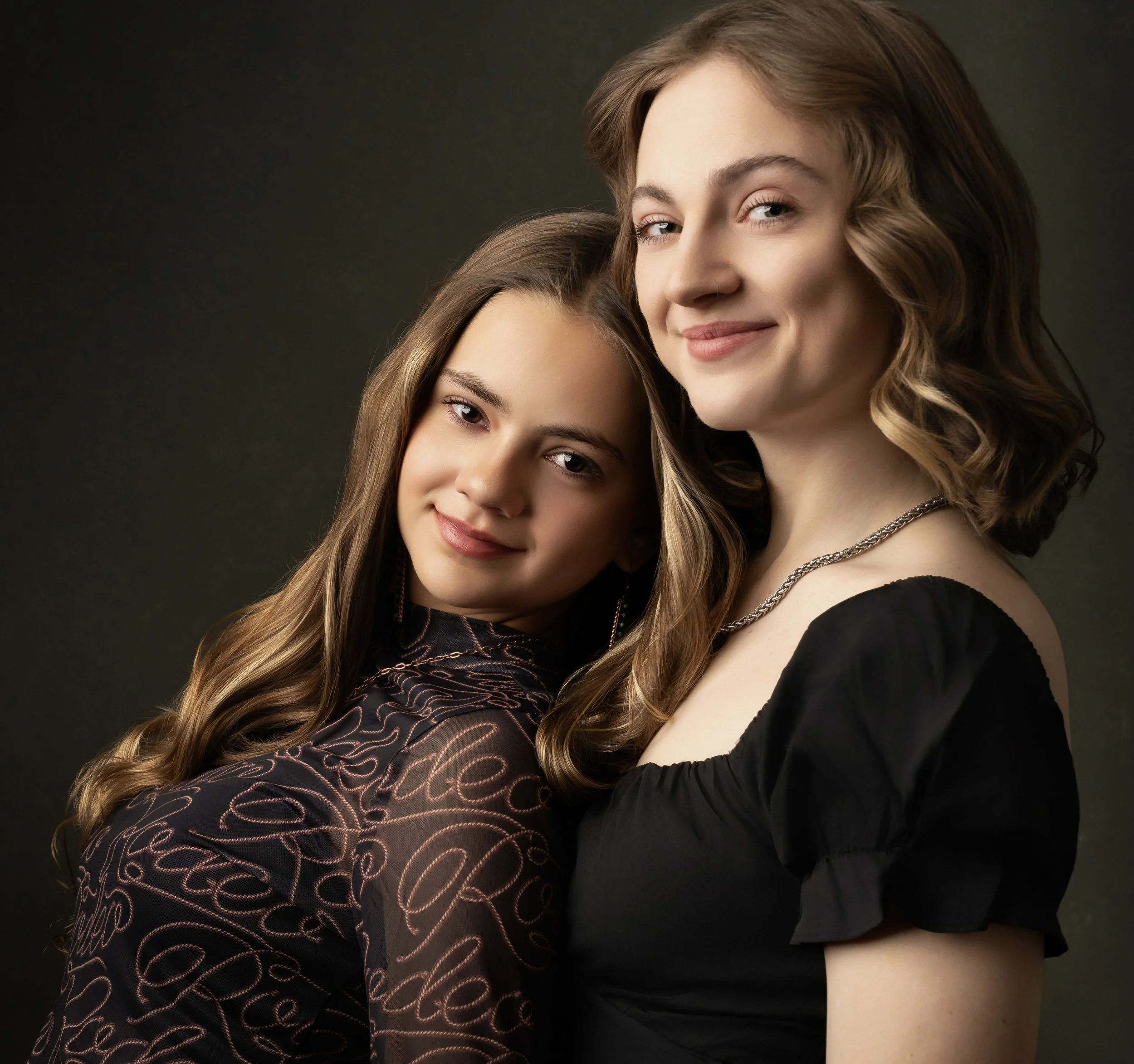 Close-up portrait of two women with long wavy hair, smiling softly, against a dark background.