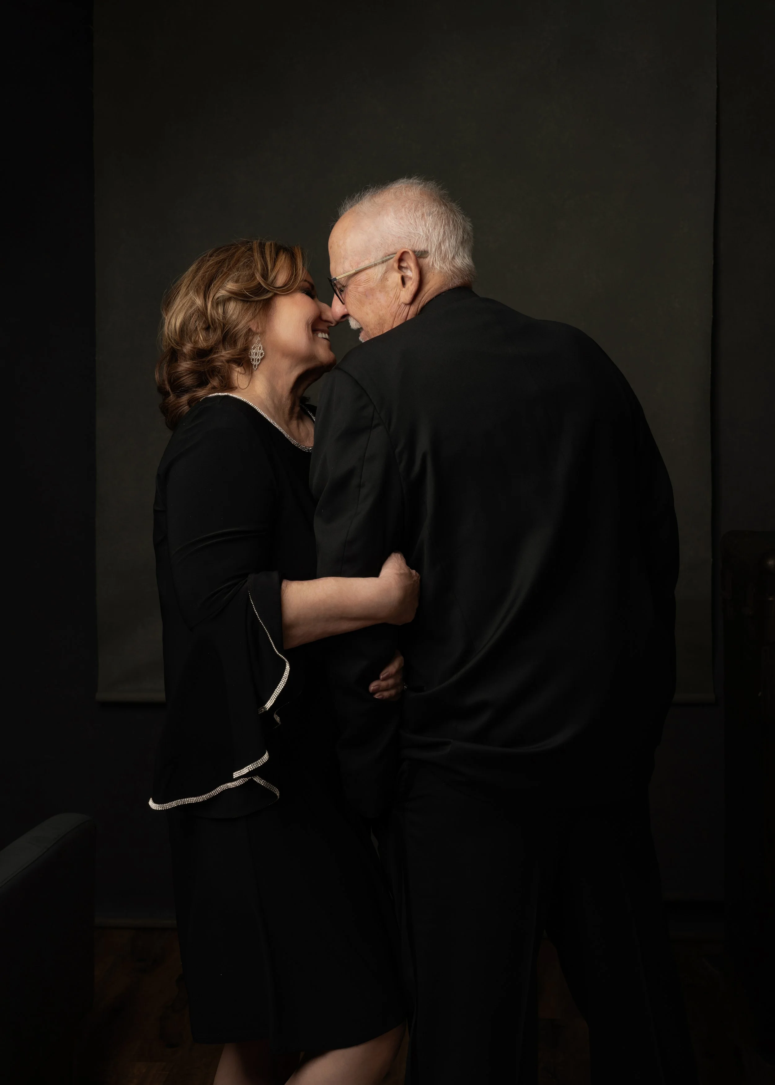 A woman and an older man are close together, smiling and touching foreheads, in a warm intimate moment. They are dressed in black, with a dark background.