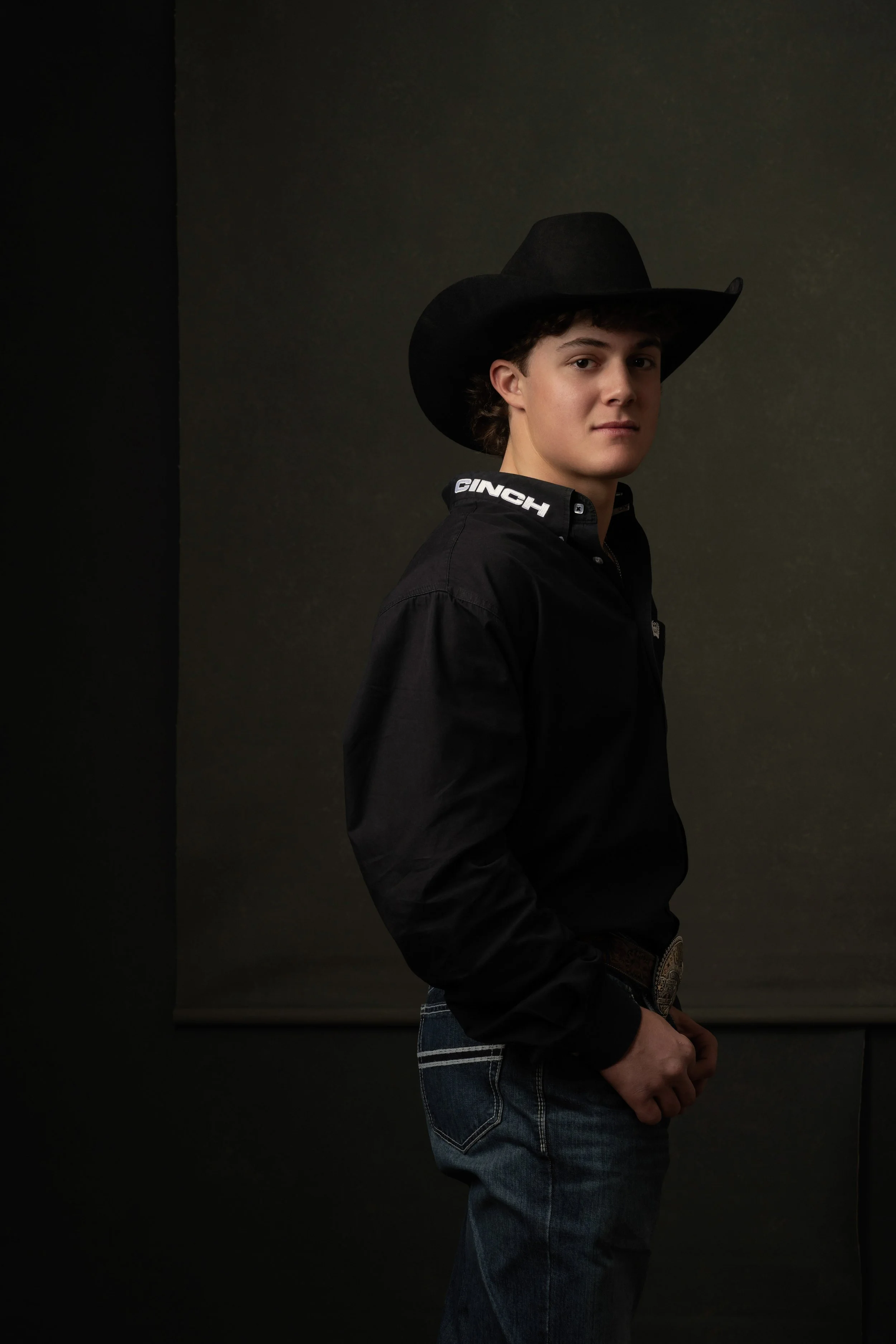 Young man wearing a black cowboy hat, black jacket with 'CINCH' logo, and jeans, standing against a dark background.