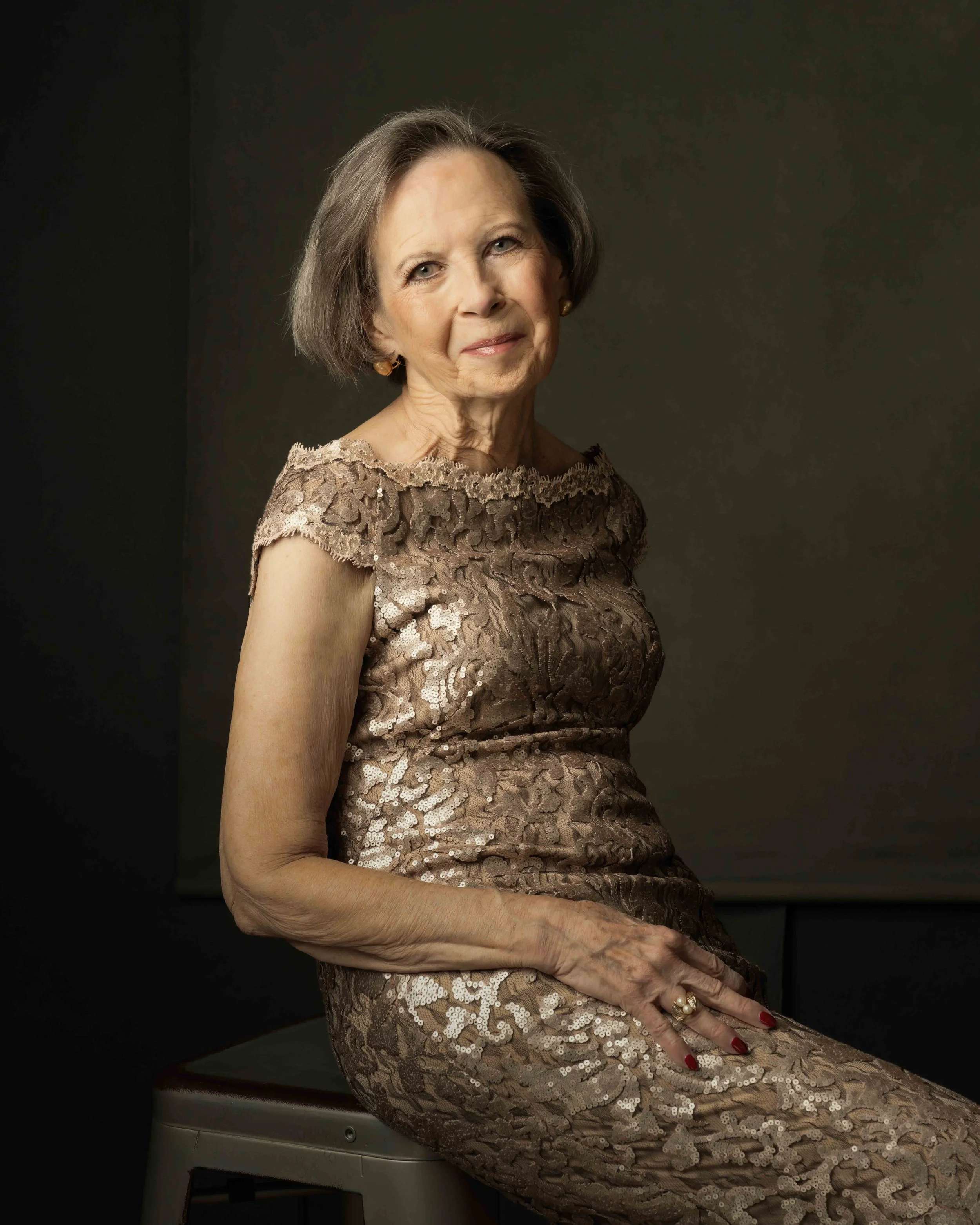 An elderly woman with short gray hair wearing a gold lace dress, sitting on a white chair, with a dark background.