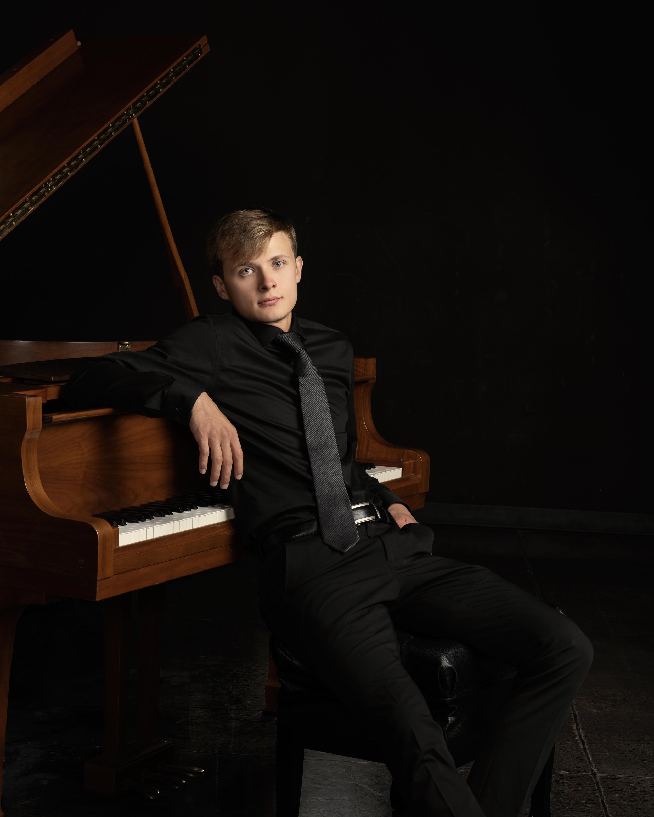 Young man with light brown hair, wearing a black shirt, gray tie, and black pants, sitting on a piano bench in front of a wooden grand piano with the lid open, against a black background.