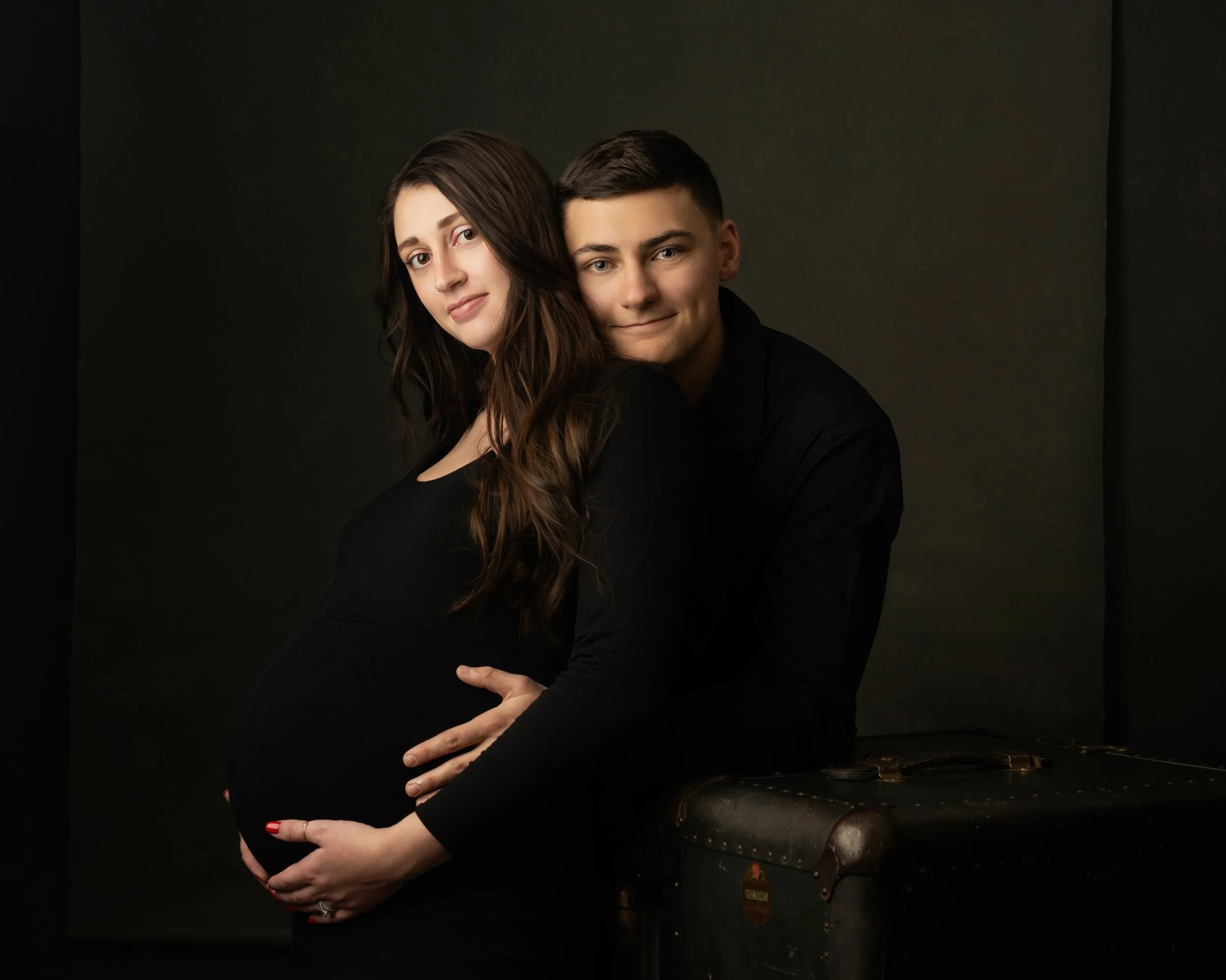 A person with dark hair and a black outfit holding a pregnant belly, standing next to a black piano against a dark background.