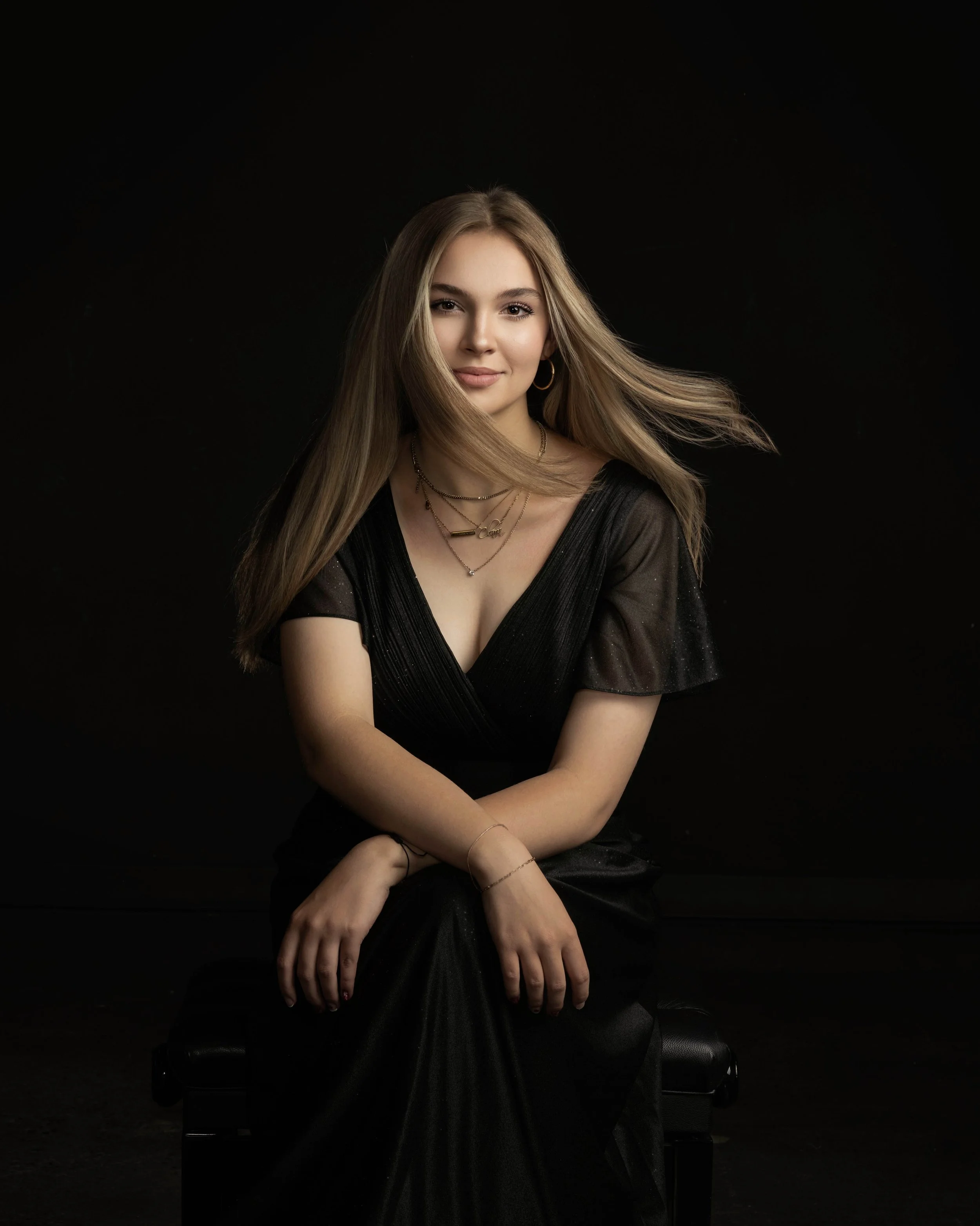 A young woman with long blonde hair sitting on a black chair against a black background. She wears a black dress, multiple layered necklaces, hoop earrings, and a bracelet. Her arms are crossed on her lap, and she has a slight smile.