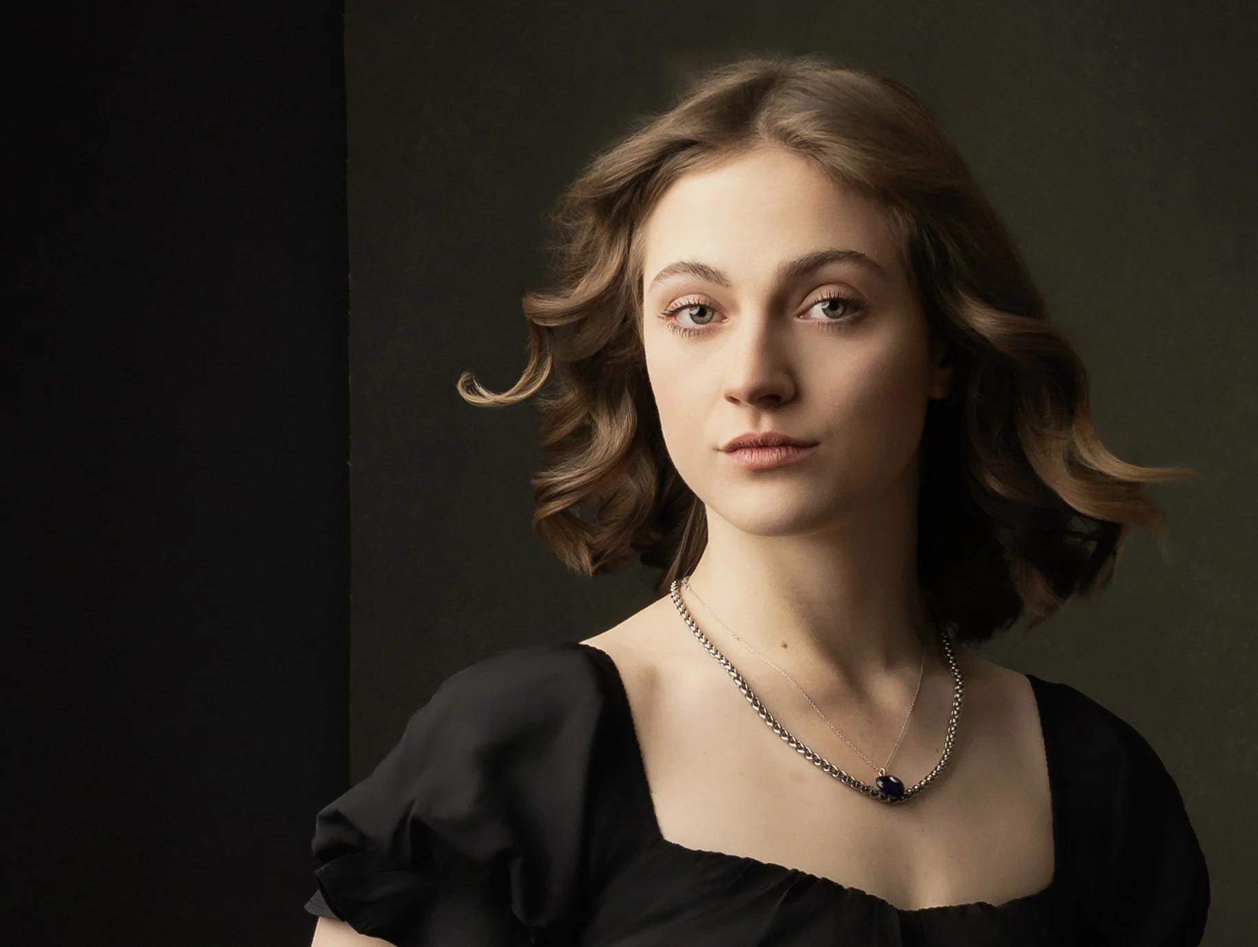A young woman with wavy brown hair, wearing a black top with puffed sleeves, layered necklaces, and makeup, standing against a dark background.