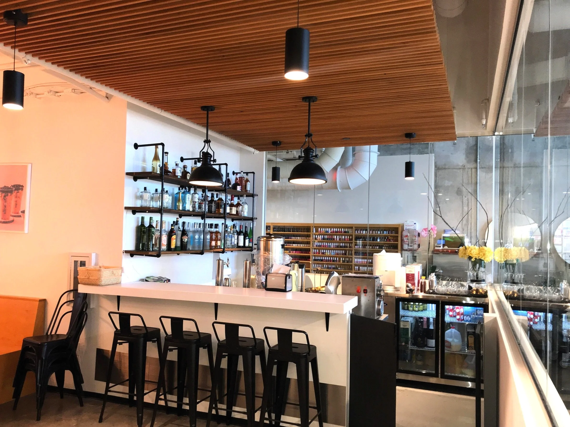 Interior of a modern cafe or bar with a white counter, black barstools, and shelves with various bottles behind the counter. There are black pendant lights hanging from a wooden ceiling and a glass wall with flowers and a view of buildings outside.